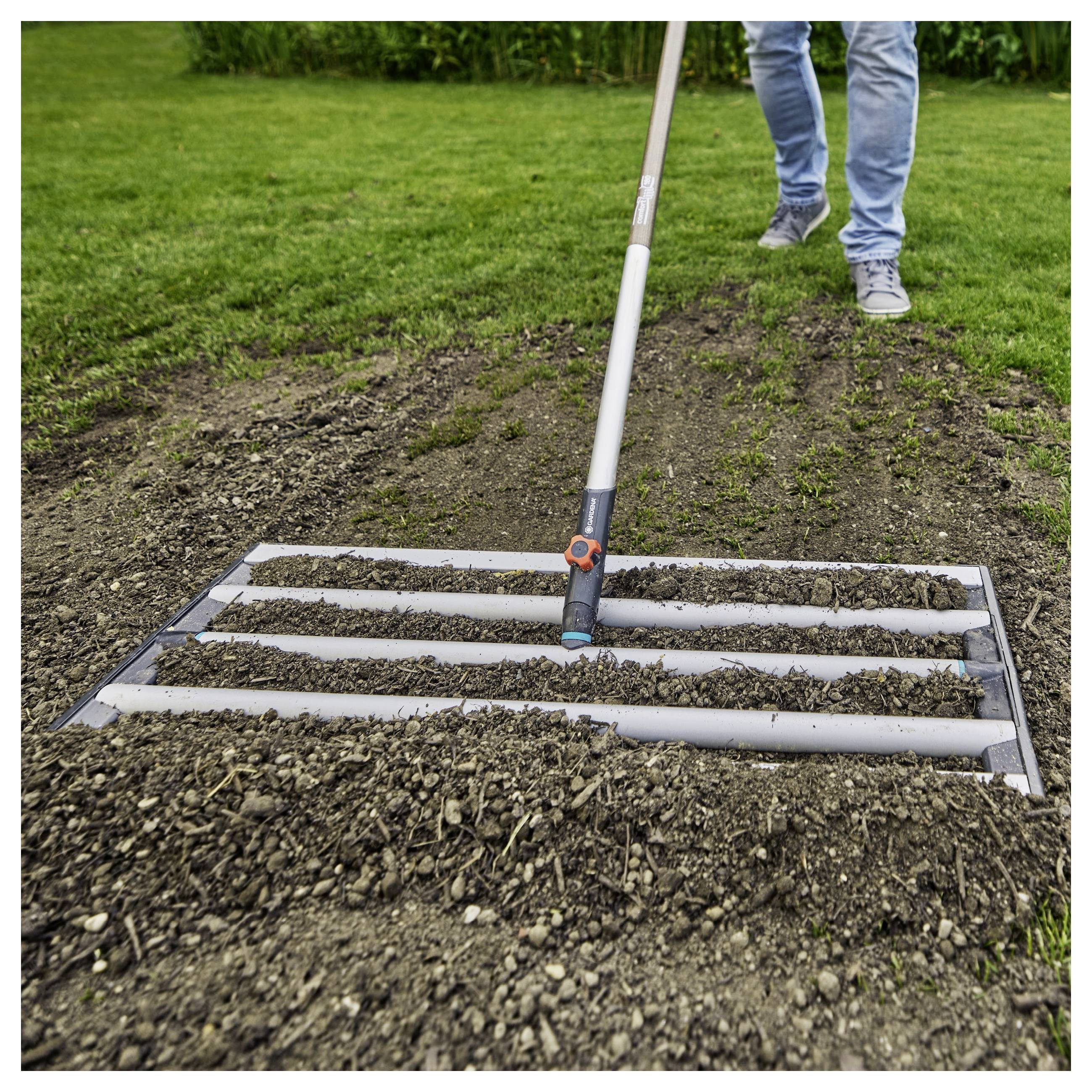 Una persona che utilizza uno strumento da giardino con barre orizzontali per livellare il terreno appena arato su un prato, suggerendo la preparazione per la semina.