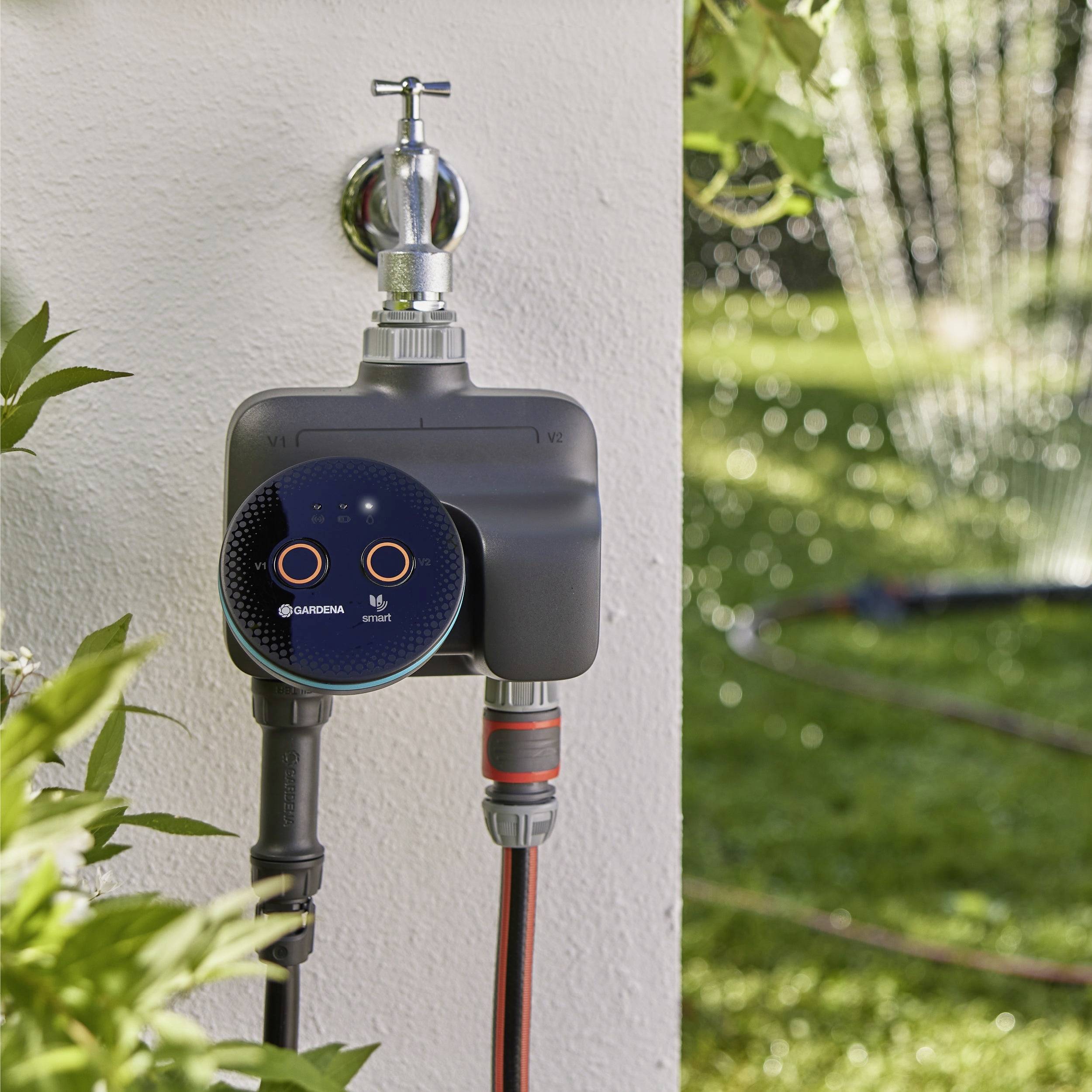 Timer automatico per acqua fissato a un rubinetto esterno, che regola gli spruzzatori del giardino. Foglie verdi e spray d'acqua sono visibili sullo sfondo.