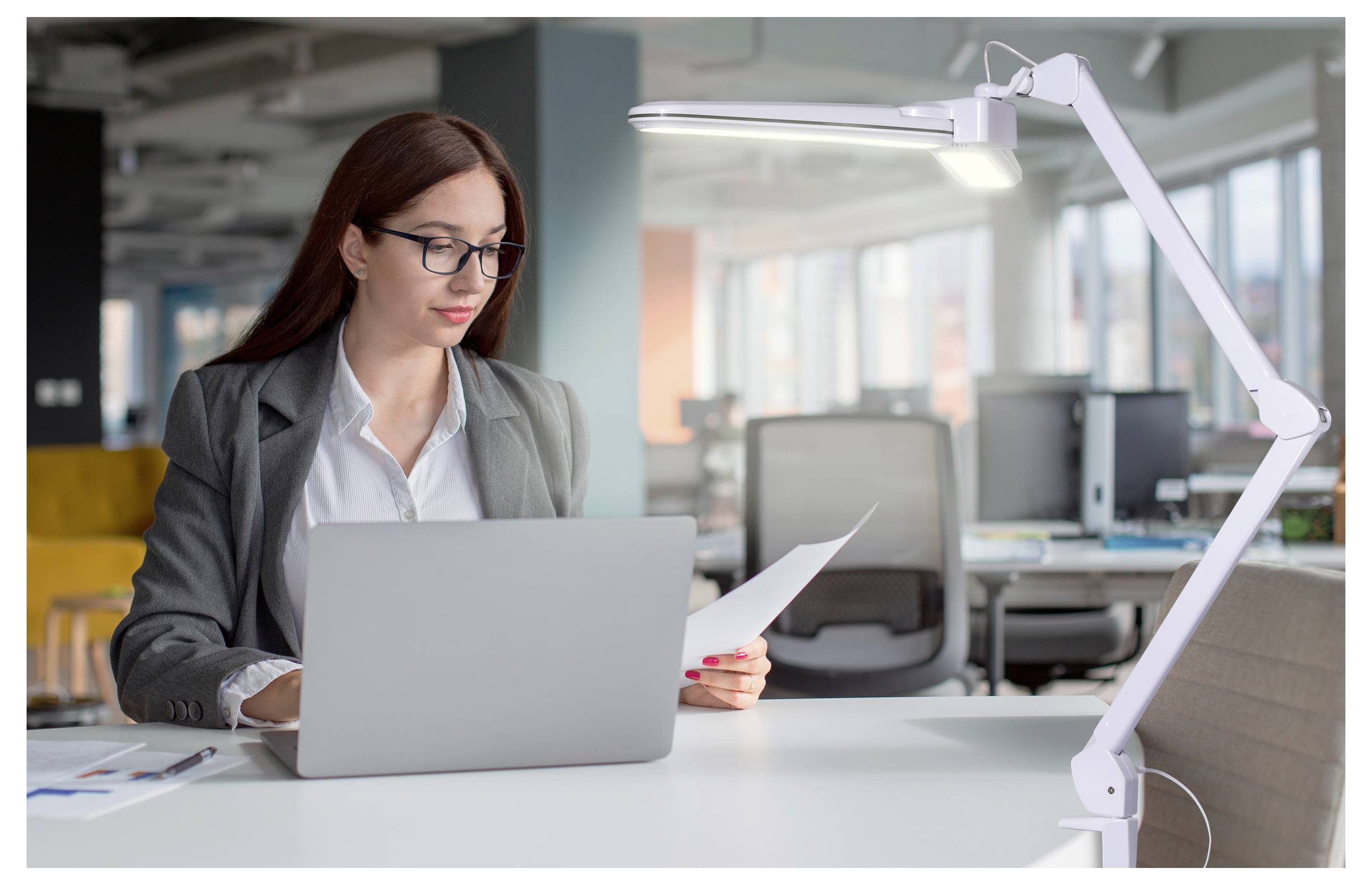 Una persona in una giacca grigia lavora al computer portatile in un moderno ambiente di ufficio, guardando un documento. Una lampada da scrivania luminosa illumina l'area di lavoro.