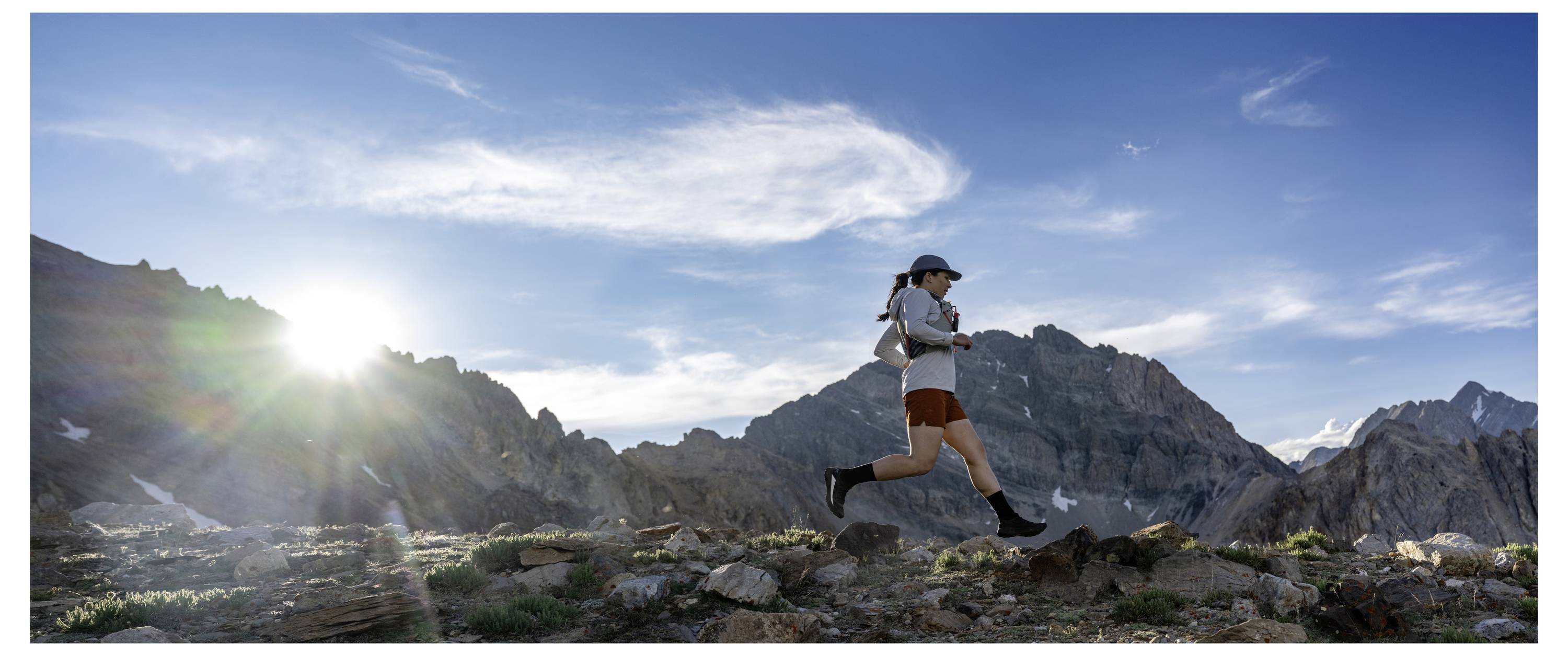 Un corridore corre lungo un sentiero di montagna sassoso, con un sole brillante e un cielo drammatico sullo sfondo, circondato da vaste e aspre vette.