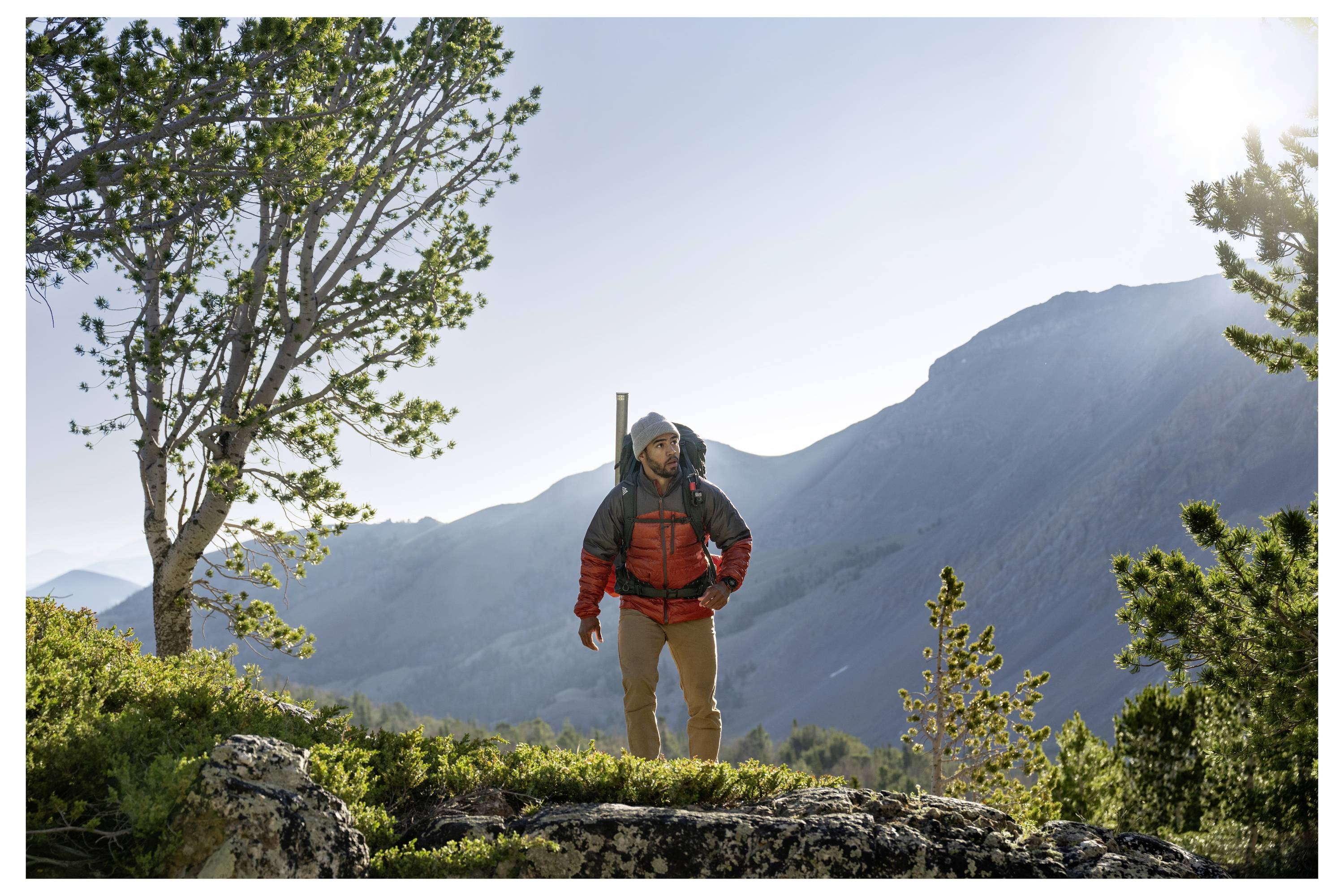 Un escursionista con una giacca rossa è in piedi su un terreno roccioso circondato da alberi, con montagne e un cielo luminoso sullo sfondo.
