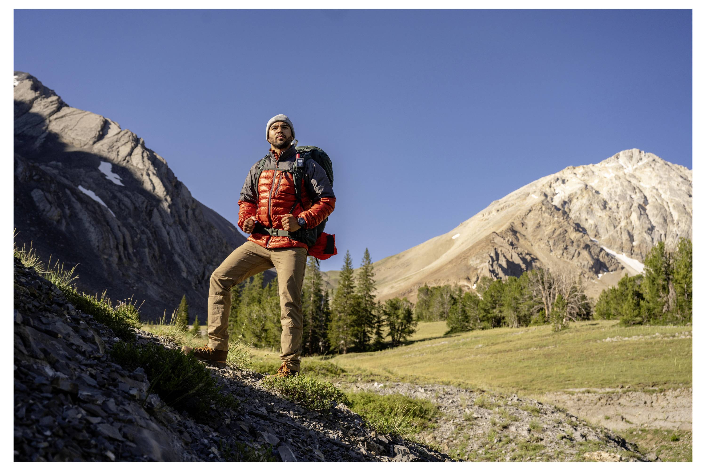 Una persona con abbigliamento da escursionismo è in piedi con sicurezza su un sentiero roccioso, con un cielo azzurro terso e maestose montagne sullo sfondo.