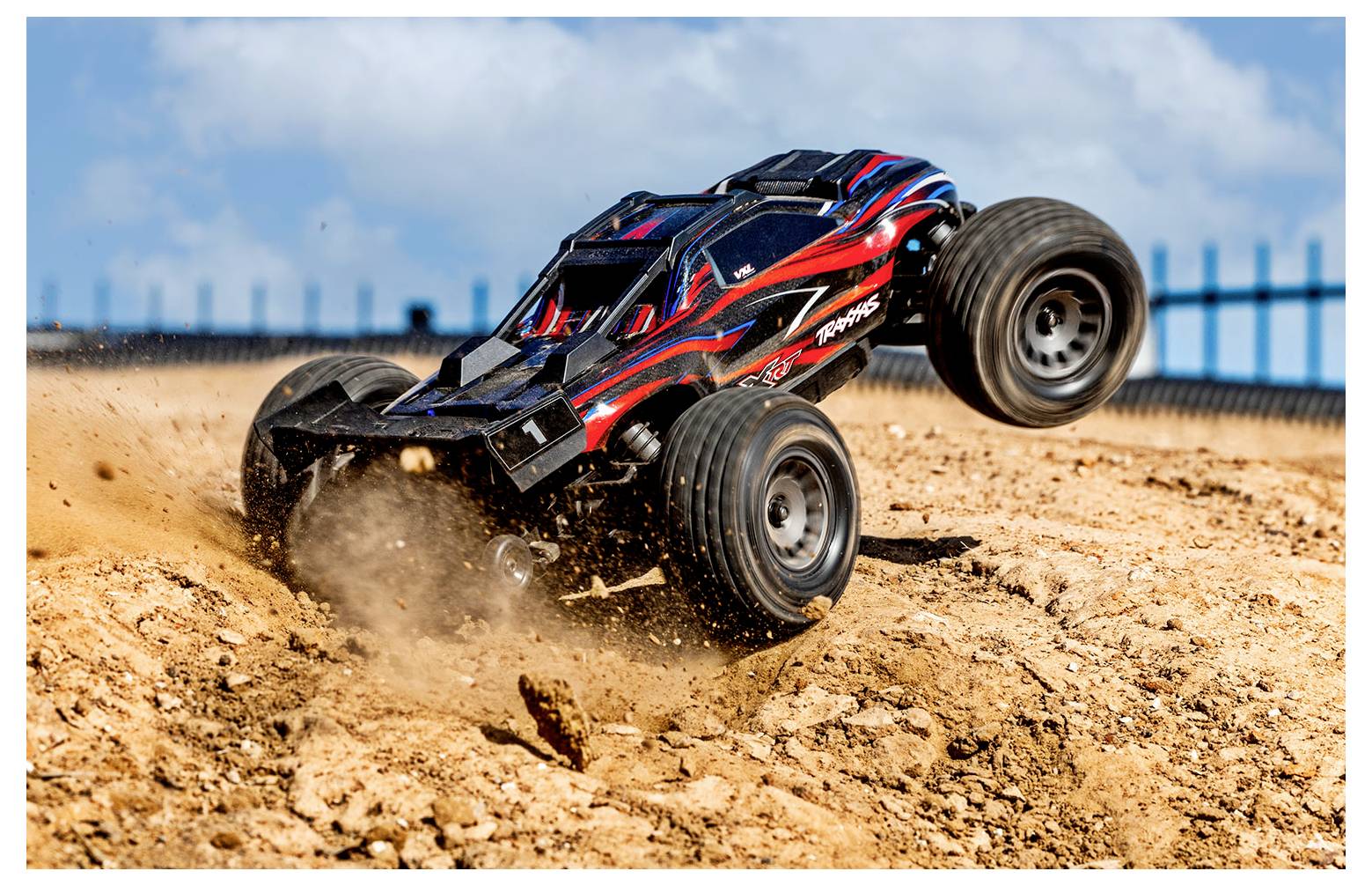 Un'auto telecomandata sta saltando sopra un cumulo di terra in un ambiente esterno, sollevando polvere sotto un cielo blu con alcune nuvole.