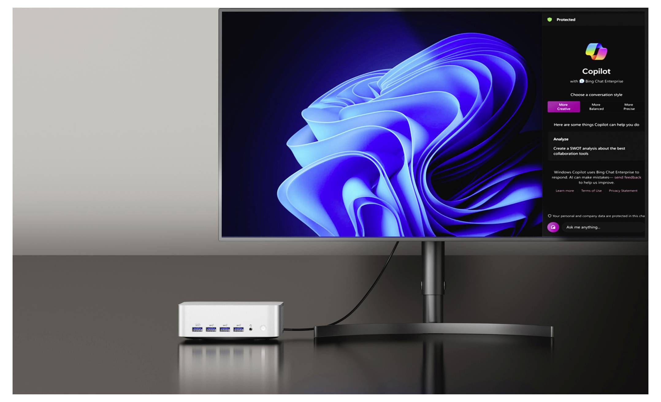 Un monitor del computer mostra un design astratto blu, con un mini PC desktop collegato tramite cavo, che visualizza un'interfaccia dell'app di intelligenza artificiale Copilot.