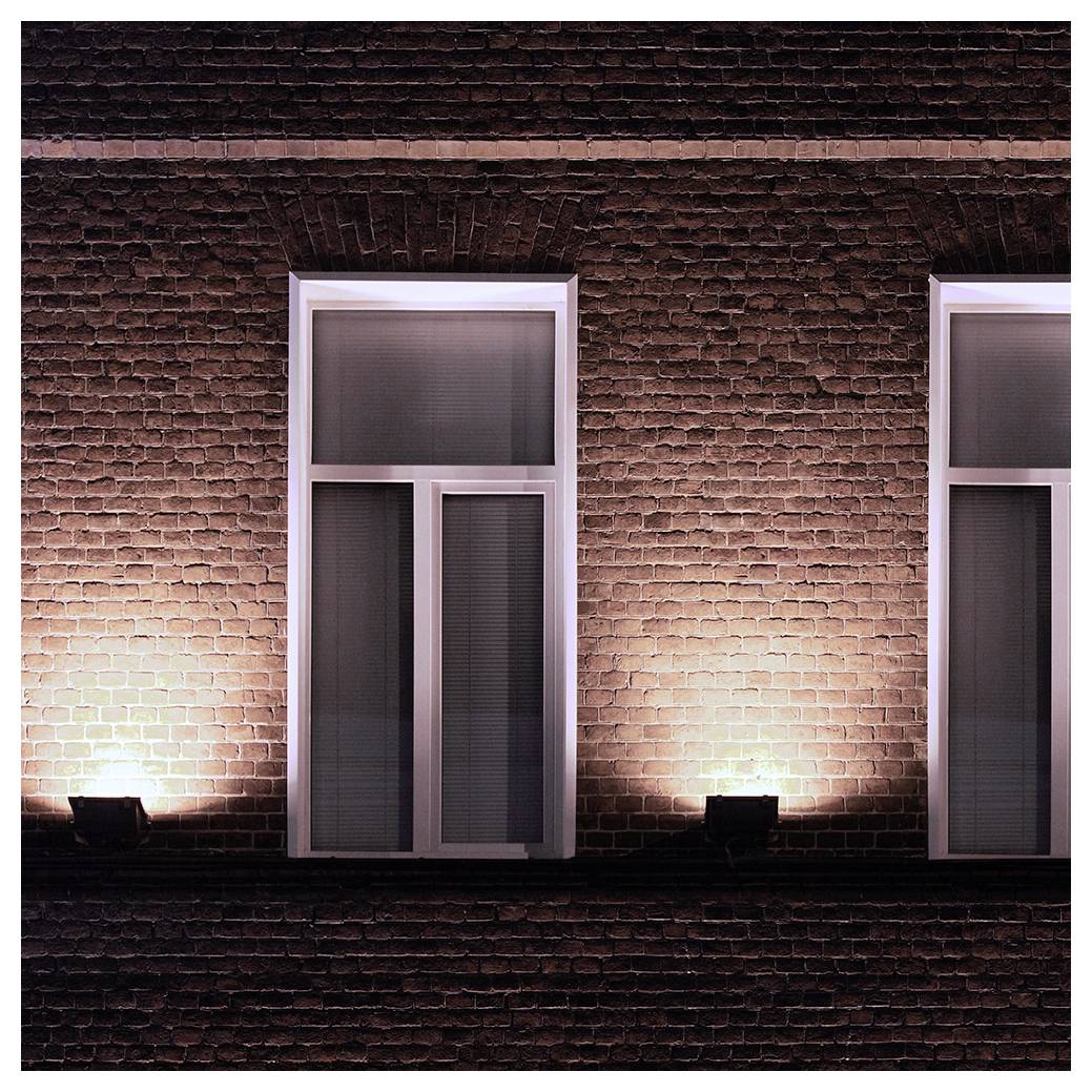 Un muro di mattoni illuminato con tre finestre verticali durante la notte, ciascuna parzialmente coperta da persiane, proietta ombre create dall'illuminazione dal basso.