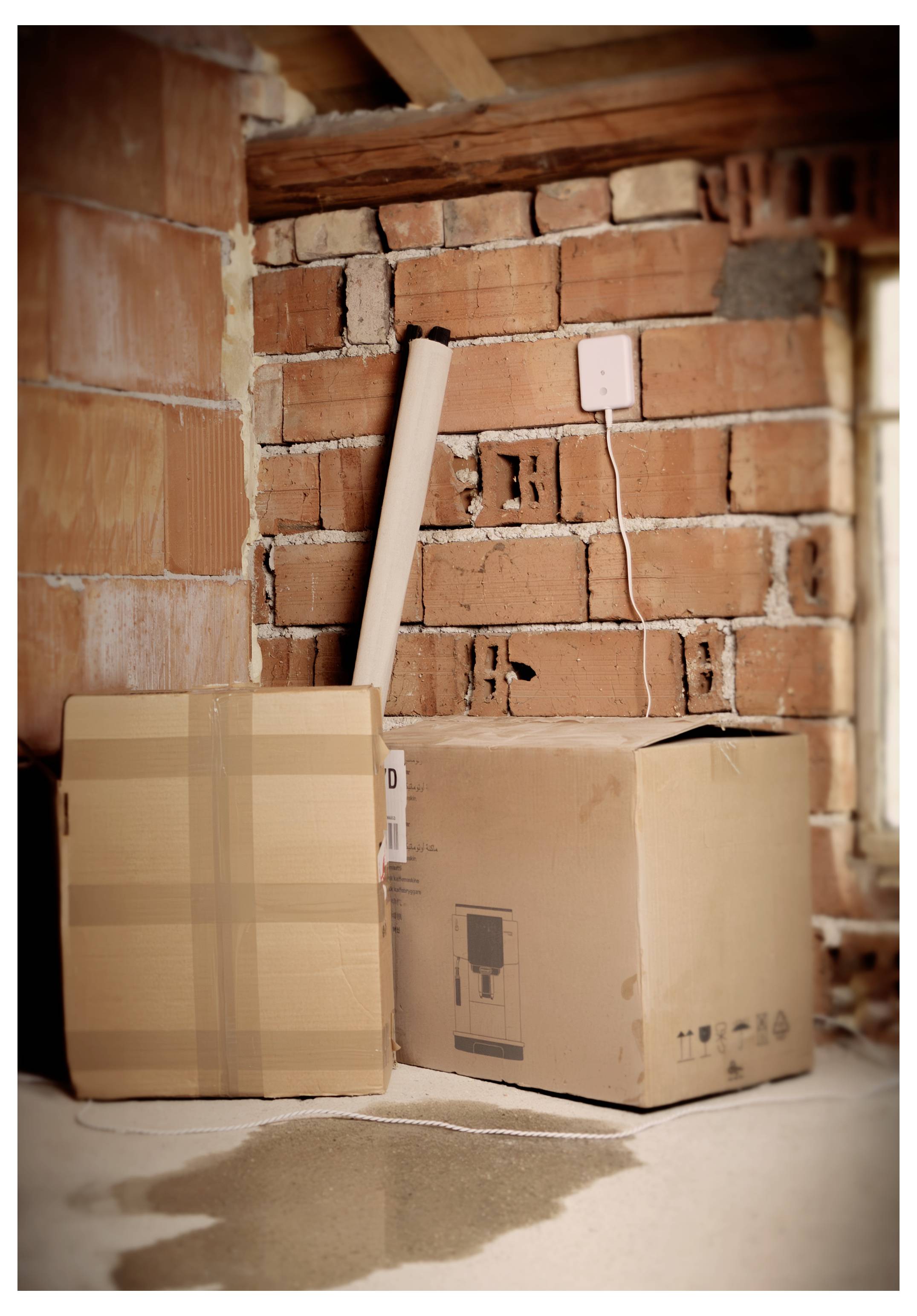 Due scatole di cartone e un tappeto arrotolato in una stanza parzialmente costruita con pareti di mattoni visibili e un pavimento in cemento.