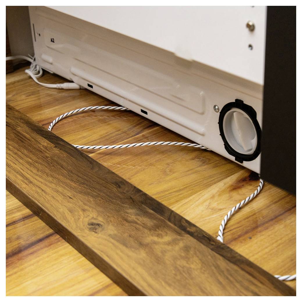 Bordo di un pavimento in legno dove incontra la base bianca di un elettrodomestico. Una presa d'aria con un tubo è parzialmente visibile, suggerendo un impianto di ventilazione.