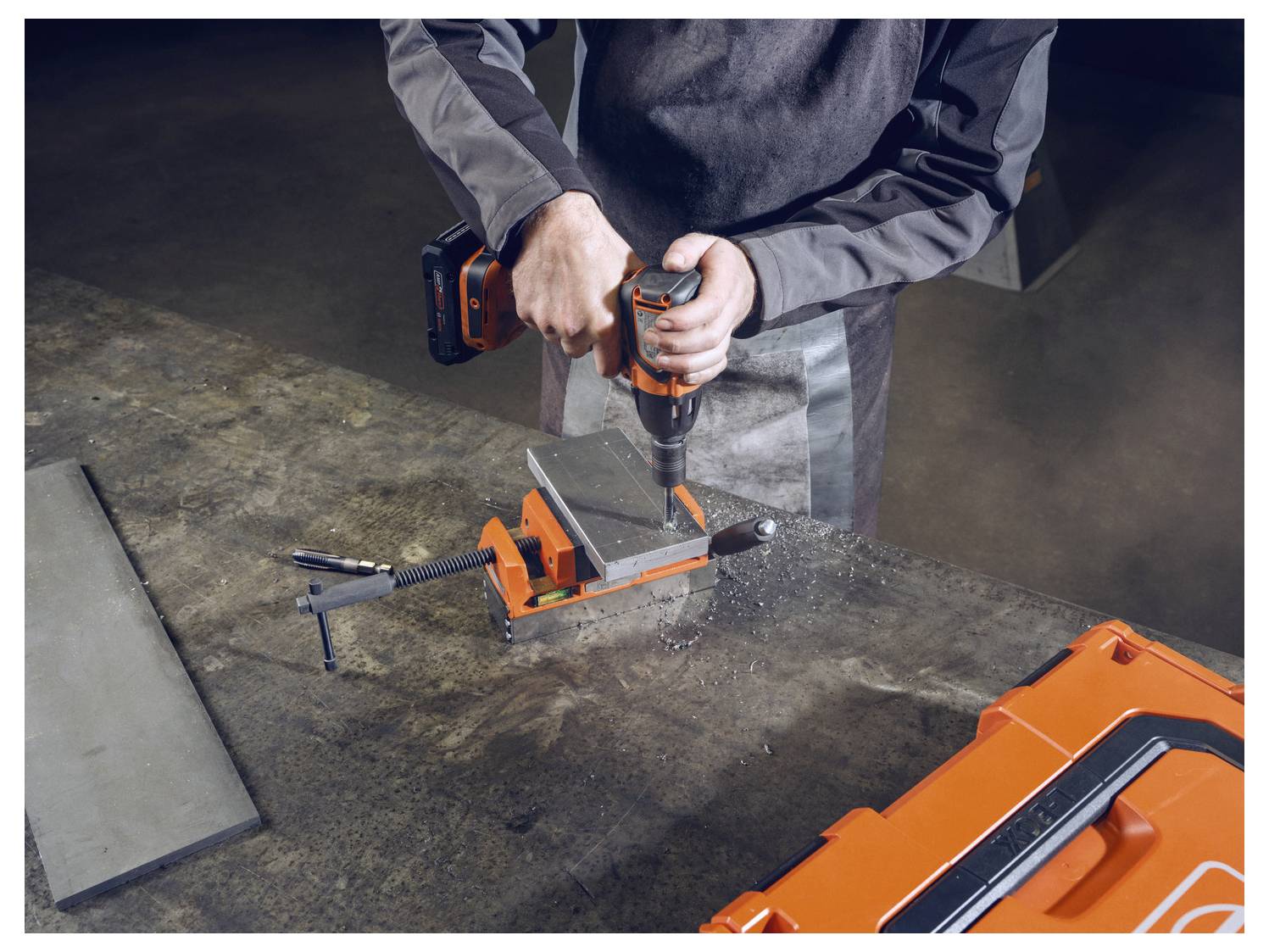 Una persona aziona una pressa trapano elettrica arancione su un tavolo di metallo, bloccando un pezzo di metallo, circondata da una cassetta degli attrezzi arancione.