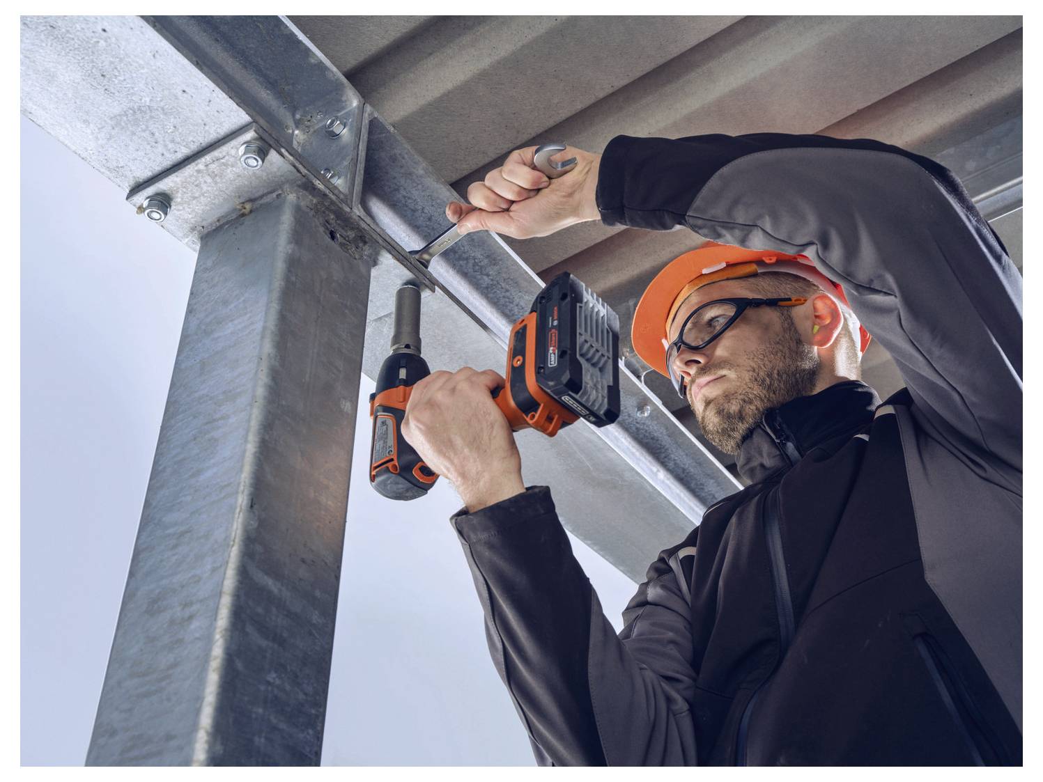 Una persona che indossa equipaggiamento di sicurezza sta utilizzando un trapano elettrico per fissare una trave metallica in un ambiente industriale.