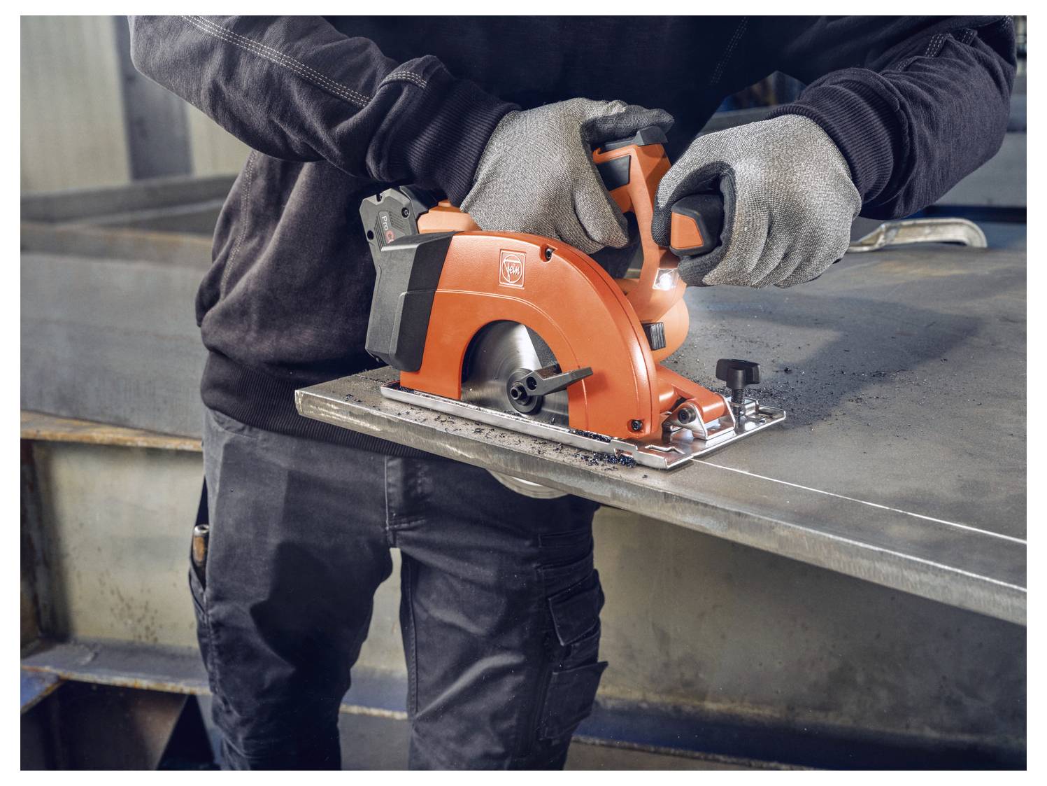 Una persona che indossa guanti utilizza una sega circolare arancione per tagliare un foglio di metallo in un ambiente industriale.