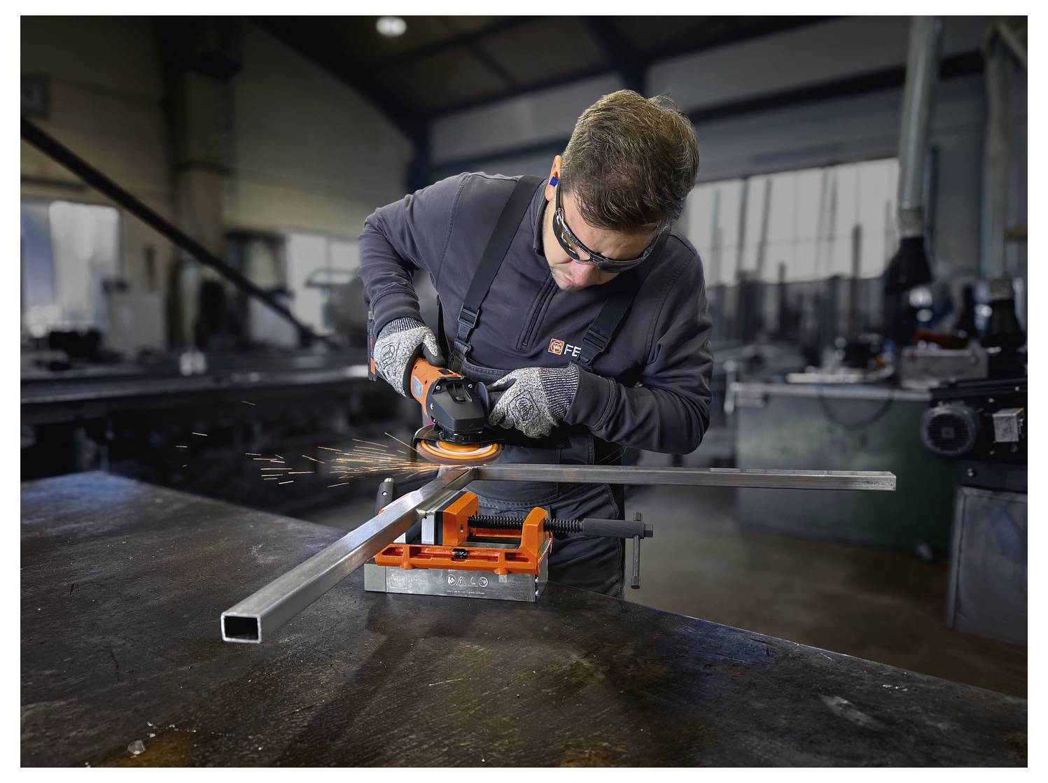 Una persona in un laboratorio che indossa equipaggiamento di sicurezza sta utilizzando una smerigliatrice angolare per tagliare un tubo quadrato di metallo fissato su un tavolo. Sono visibili scintille.