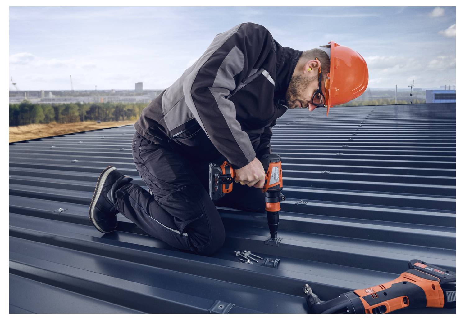 Una persona che indossa equipaggiamento protettivo utilizza un trapano a batteria su un tetto in metallo, con uno skyline della città visibile sullo sfondo.