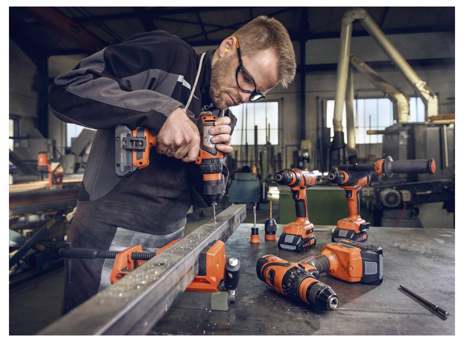 Un uomo in un laboratorio usa un trapano a percussione su una trave metallica, circondato da vari utensili elettrici su un tavolo, evidenziando il lavoro industriale.
