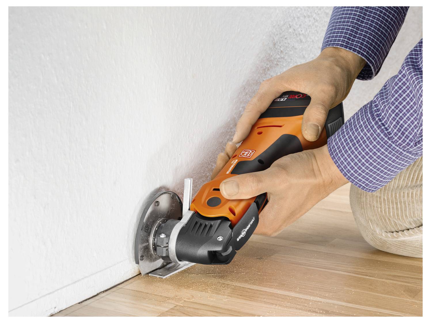 Una persona usa un attrezzo elettrico arancione per tagliare un pavimento in legno lungo un muro bianco.
