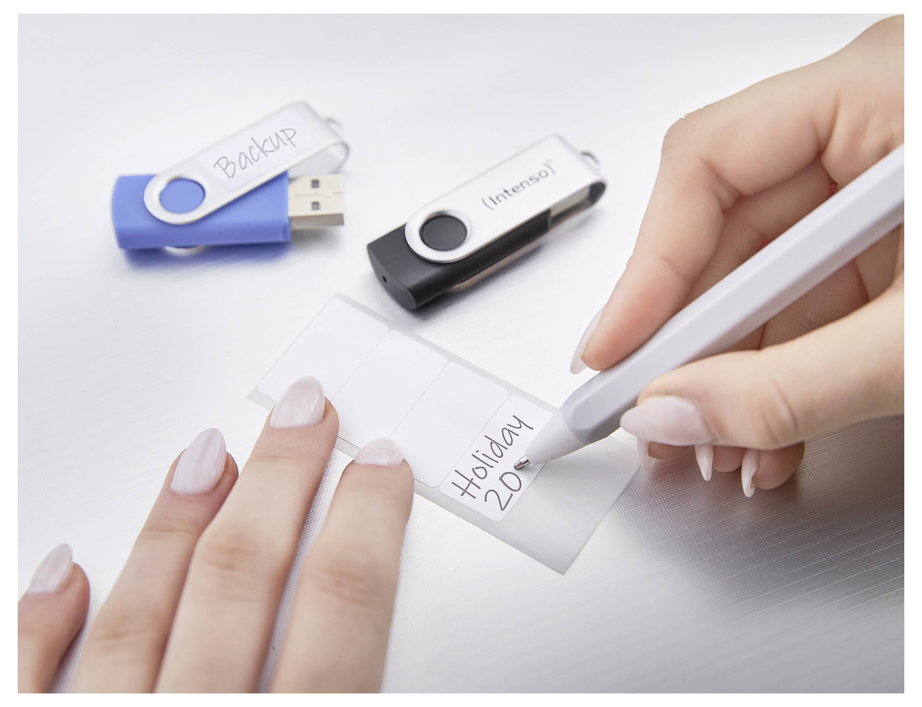 Una mano scrive "Holiday 2020" su un'etichetta mentre due chiavette USB etichettate "Backup" e "Intenso" si trovano sul tavolo.