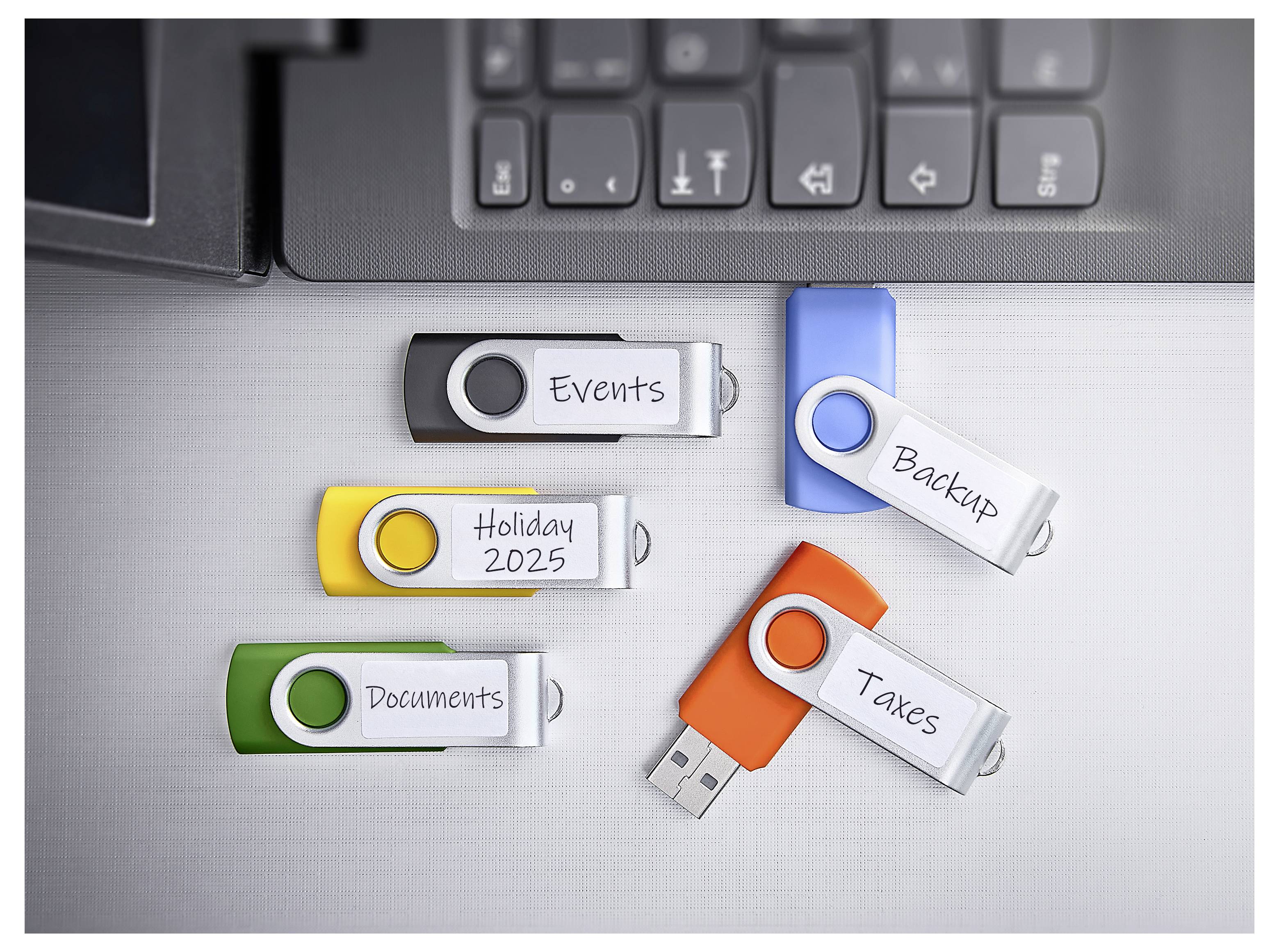 Chiavette USB etichettate 'Events', 'Holiday 2025', 'Documents', 'Backup' e 'Taxes' sono sparse vicino a una tastiera di laptop, indicando un'organizzazione dei dati.