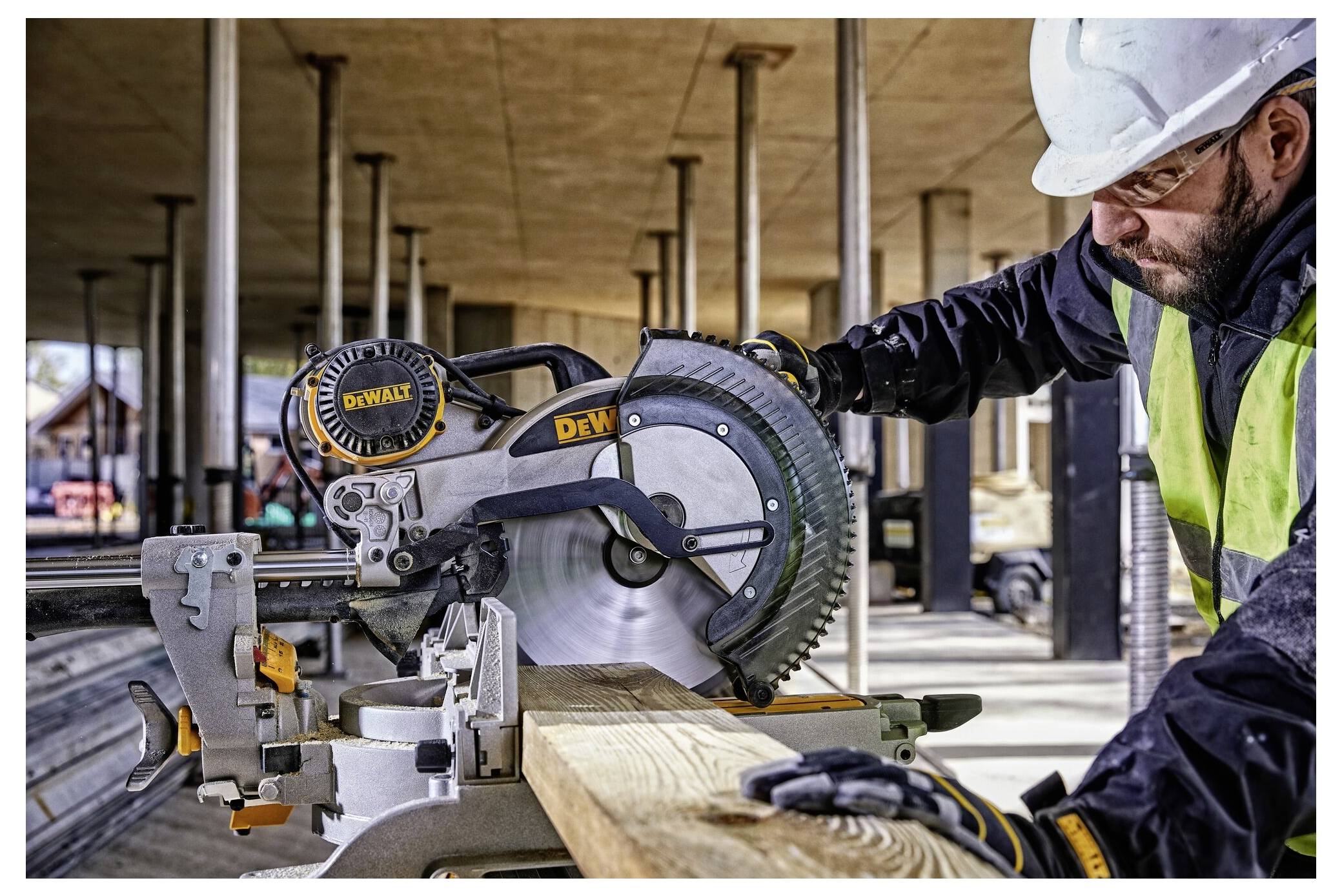 Un operaio edile con equipaggiamento di sicurezza aziona una sega circolare che taglia una tavola di legno presso un cantiere edile con colonne in calcestruzzo.