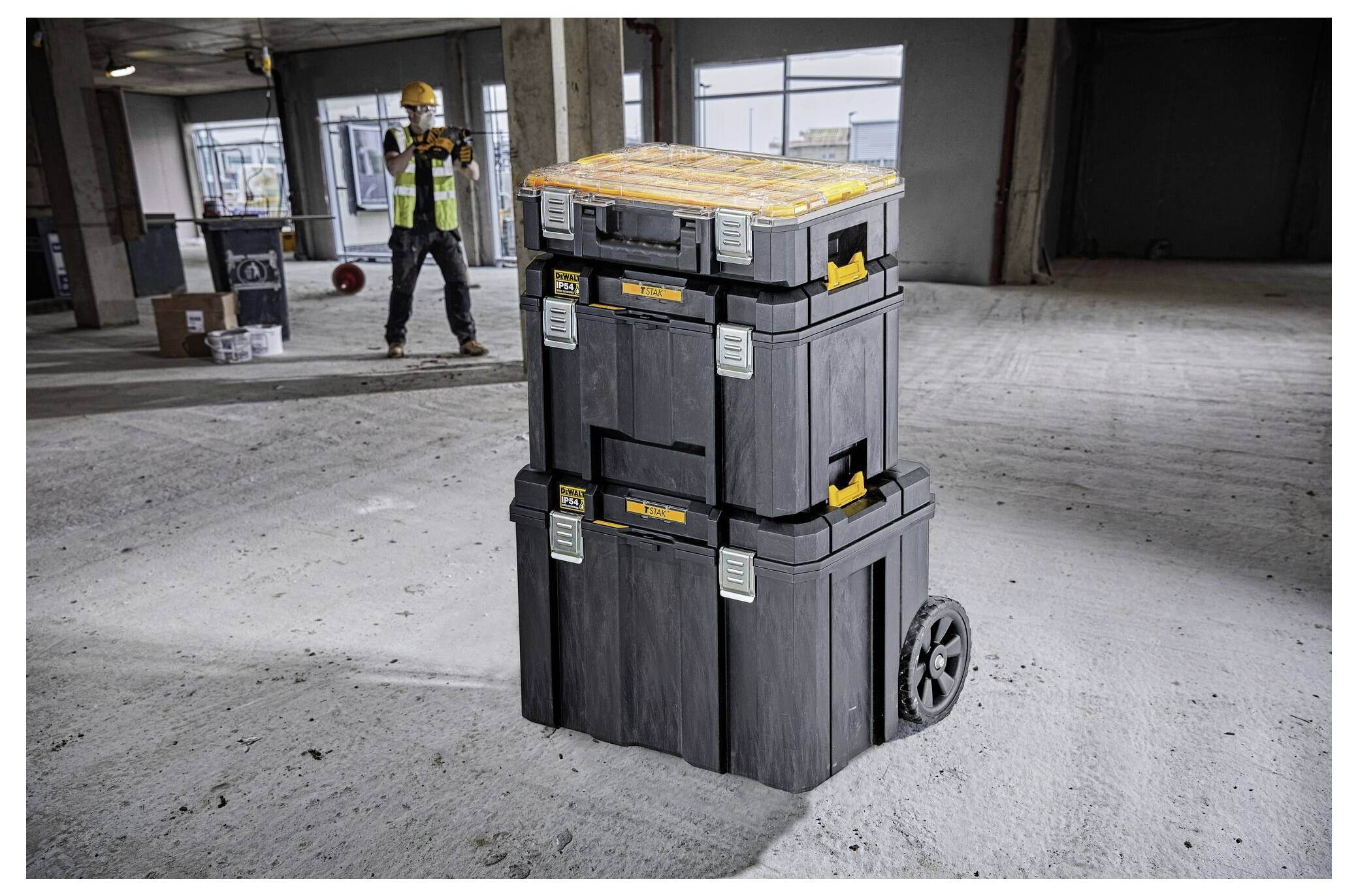 Una pila di tre scatole di stoccaggio nere con accenti gialli su ruote in un cantiere. Un operaio sullo sfondo scatta una foto.