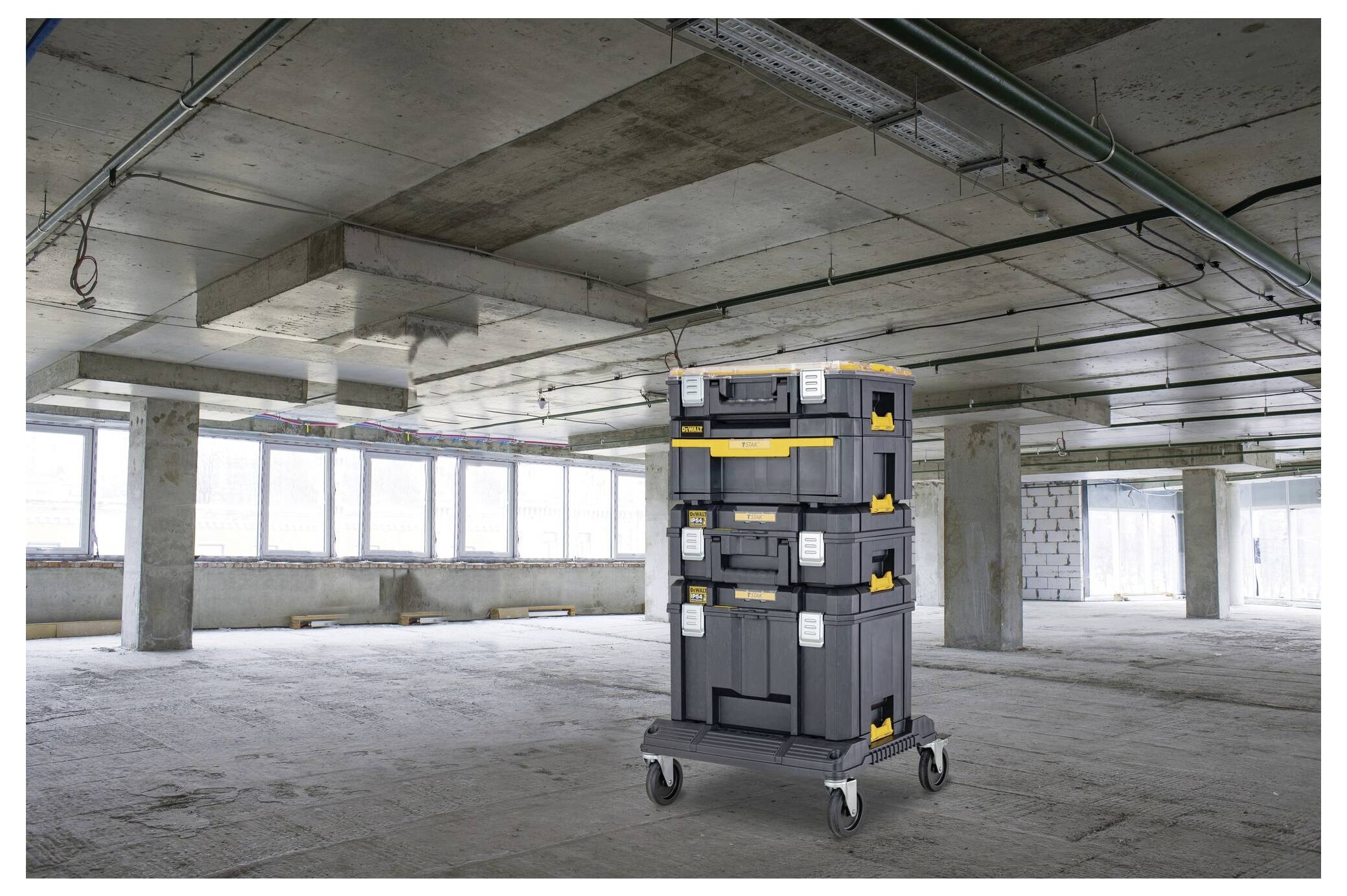 Tre scatole di stoccaggio impilate su ruote in una stanza con pavimento in cemento non rifinito, grandi finestre, tubi e pareti non rifinite.