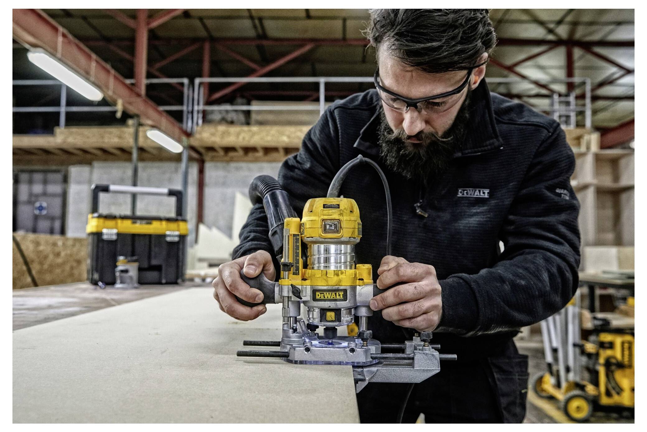 Un uomo che indossa occhiali di sicurezza utilizza un attrezzo router giallo su una tavola di legno in un ambiente di officina industriale.