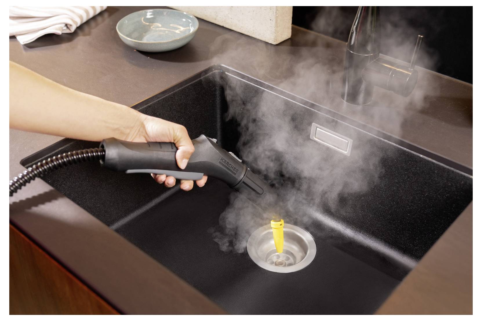 Una mano che utilizza un pulitore a vapore per disinfettare un lavello da cucina, con vapore visibilmente che sale.