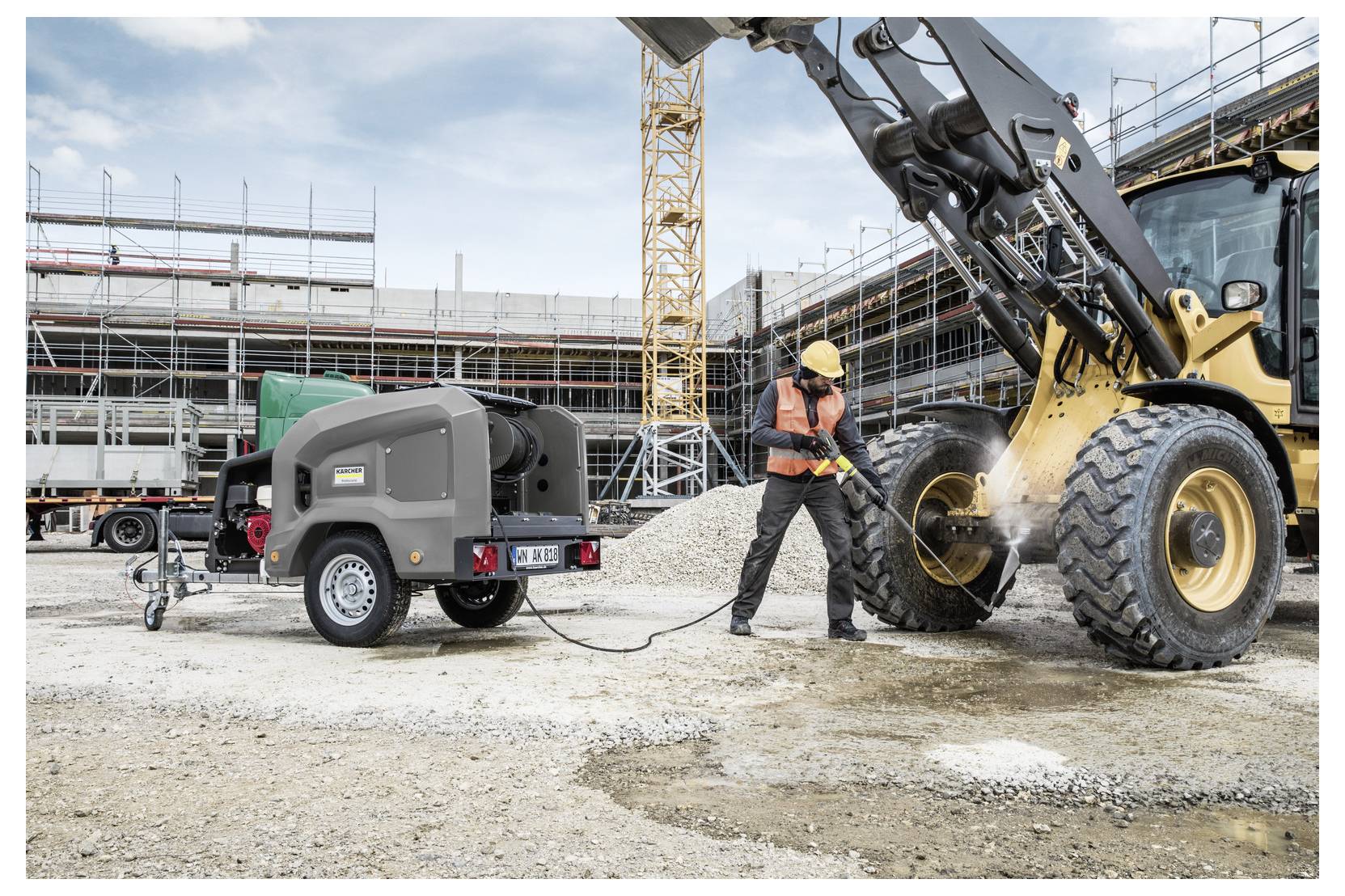 Un operaio edile utilizza un'idropulitrice ad alta pressione, collegata a un compressore d'aria portatile, per pulire il pneumatico di un grande veicolo in un vivace cantiere.