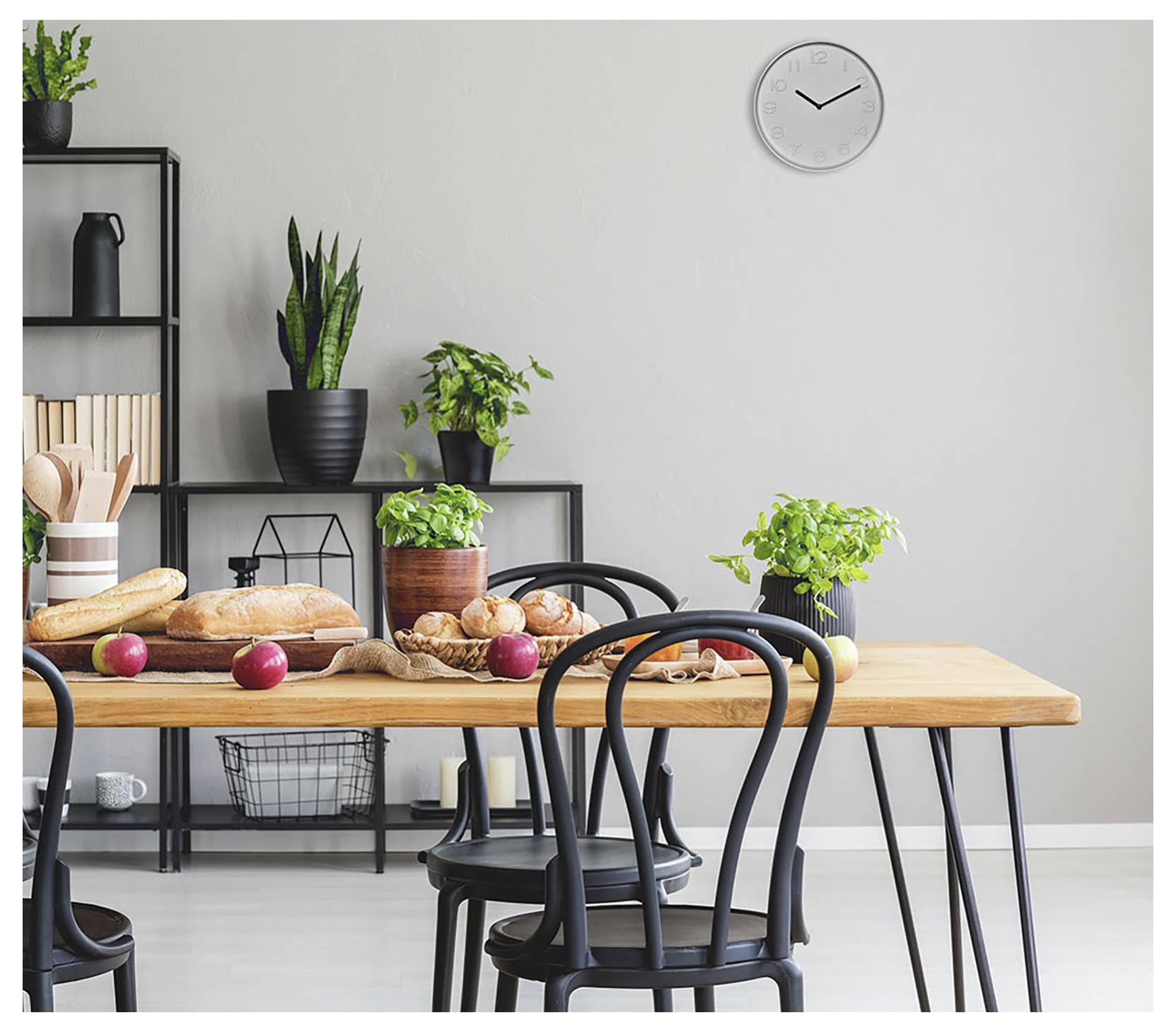 Una moderna sala da pranzo con un tavolo in legno apparecchiato con pane, mele e piante. Sedie nere circondano il tavolo. Un orologio e scaffali sono visibili.