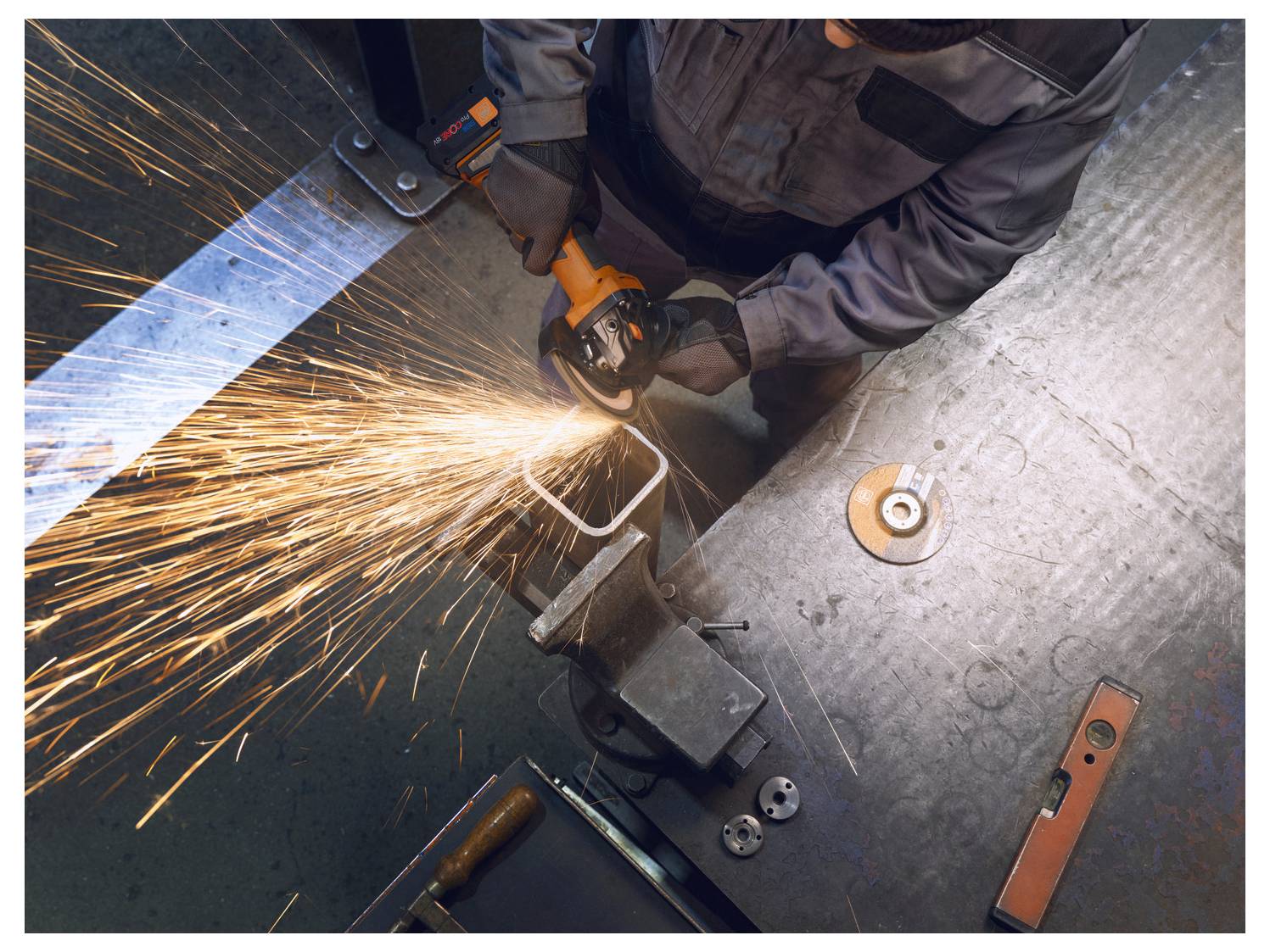 Una persona utilizza un utensile elettrico per tagliare il metallo, creando scintille luminose. La scena si svolge in un'officina con vari attrezzi e attrezzature visibili.