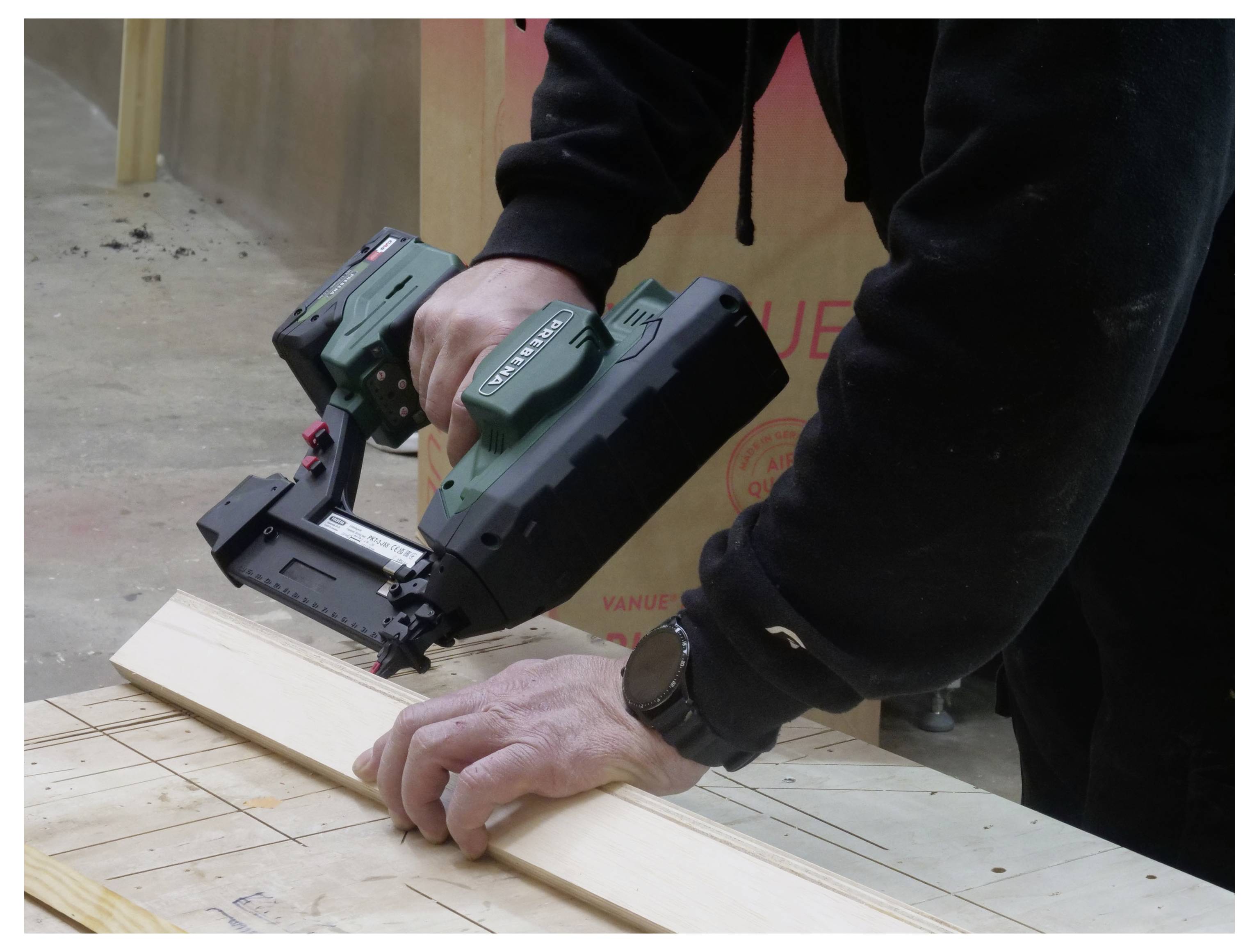 Una persona in una felpa con cappuccio nera utilizza una pistola chiodatrice elettrica verde per fissare una tavola di legno su un banco da lavoro, concentrandosi intensamente sul compito.