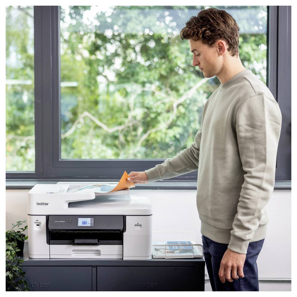 Un uomo gestisce una stampante, inserendo carta arancione nello scanner. La scena è ambientata in una stanza luminosa con una grande finestra e del verde all'esterno.