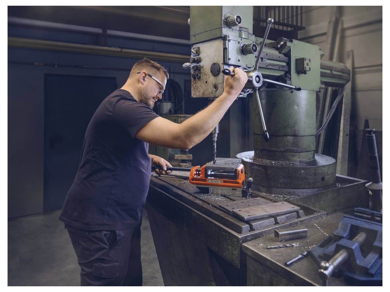 Una persona aziona una fresatrice in un'officina, regolando un componente sul tavolo. Vari attrezzi e apparecchiature sono visibili intorno.