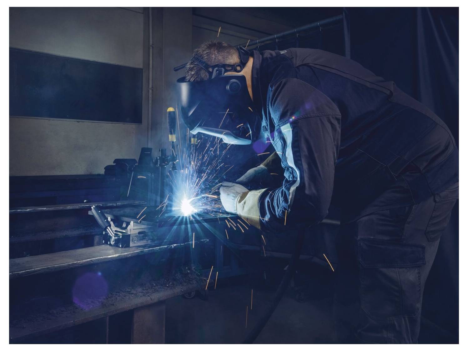 Una persona che indossa un equipaggiamento di protezione salda il metallo, creando scintille luminose in un'officina scarsamente illuminata.