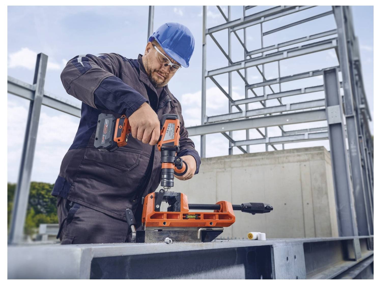 Un operaio edile in equipaggiamento di sicurezza perfora travi metalliche in un cantiere. Una struttura d'acciaio parzialmente costruita è visibile.