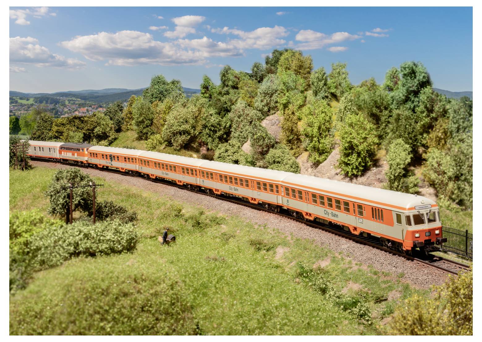 Un lungo treno arancione viaggia attraverso un paesaggio suggestivo e erboso con colline ondulate e alberi sotto un cielo blu con nuvole sparse.