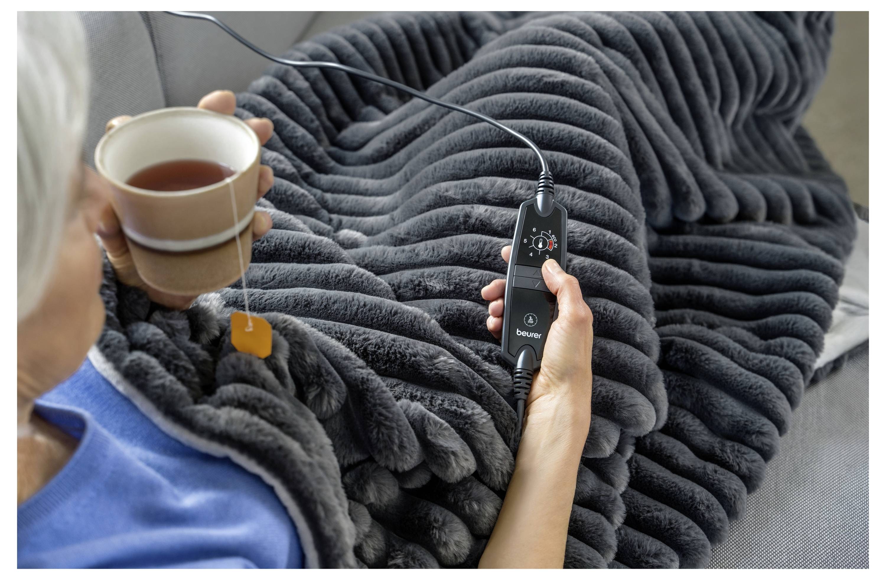 Persona avvolta in una coperta in morbido grigio, che tiene una bevanda calda e un telecomando, possibilmente regolando le impostazioni di temperatura della coperta.
