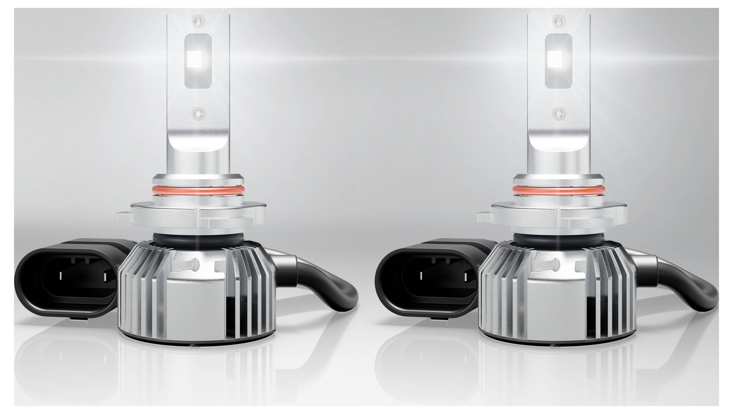 Due lampadine per fari LED con alette di raffreddamento e connettori di alimentazione, posizionate su uno sfondo riflettente, che emettono una luce soffusa.