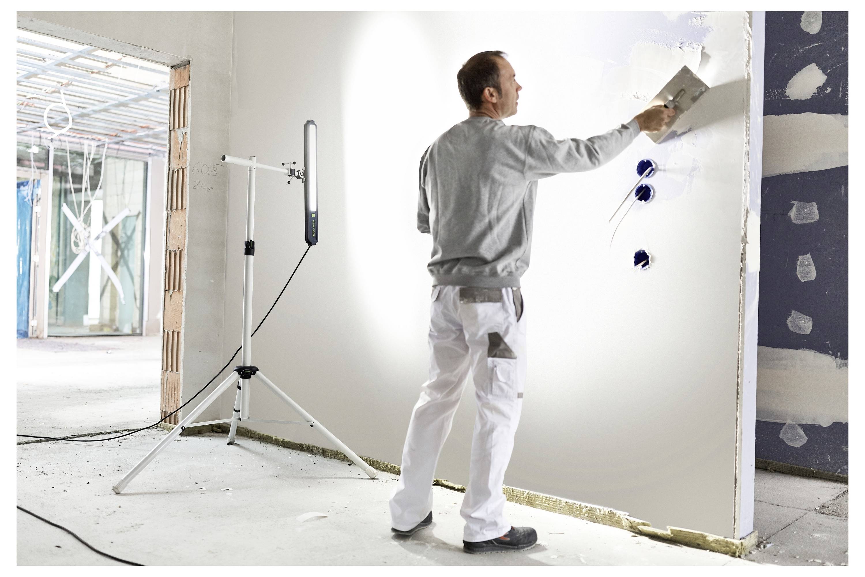 Un operaio in pantaloni bianchi e camicia grigia sta livellando l'intonaco su un muro parzialmente costruito con una cazzuola in un edificio in costruzione.