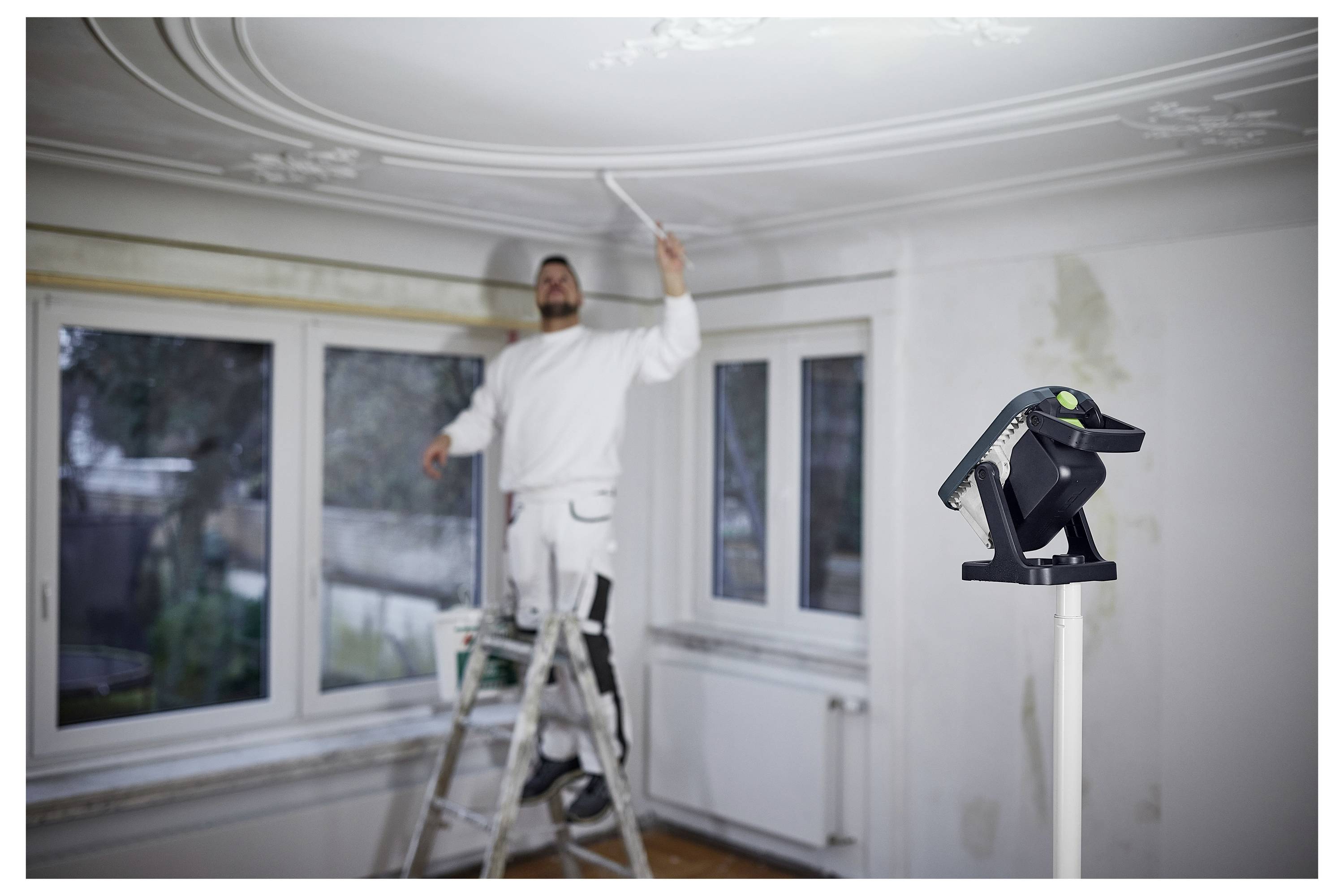 Una persona sta in piedi su una scala mentre dipinge un soffitto in una stanza con grandi finestre. Uno strumento di livello laser è posizionato in primo piano.