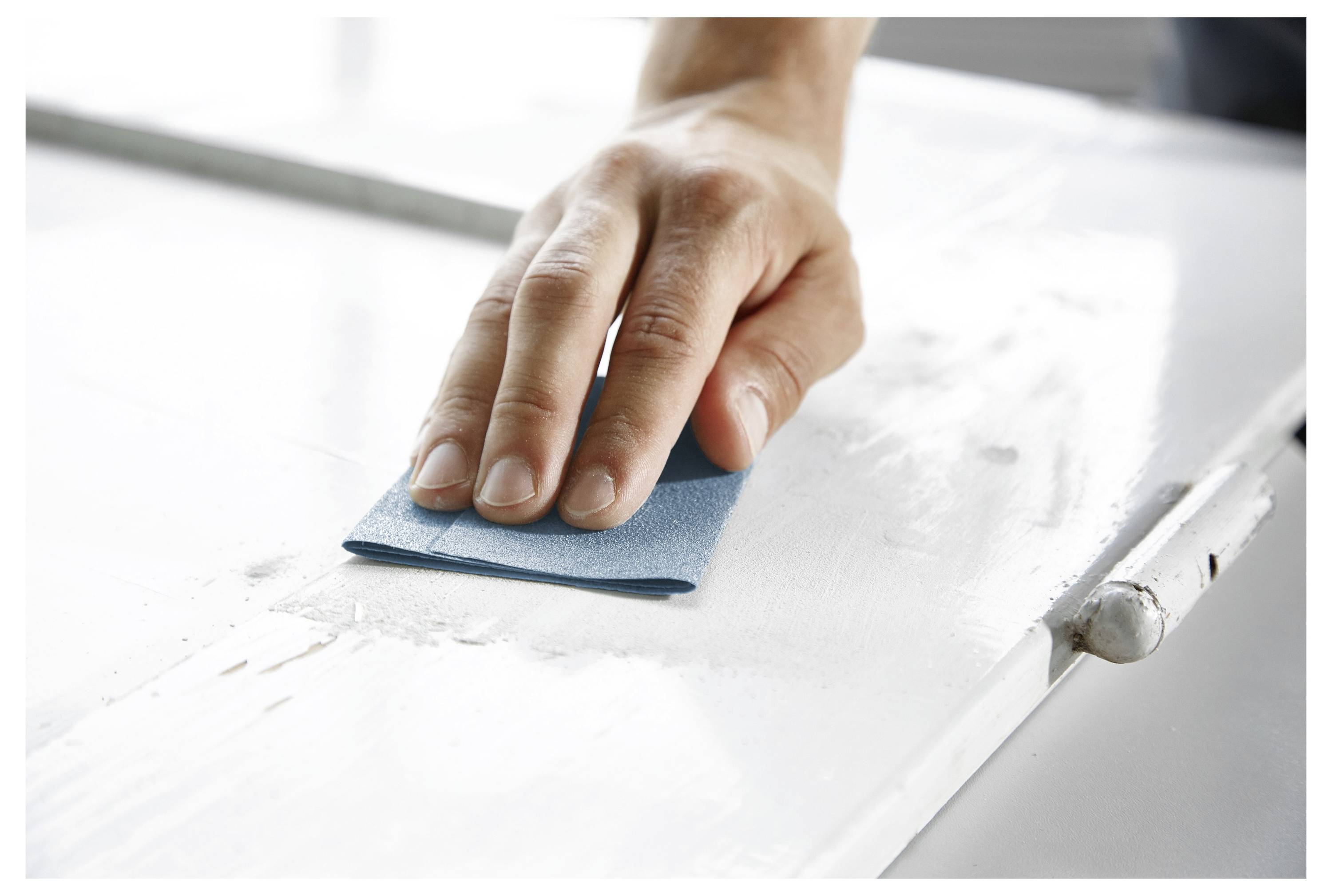Una mano che carteggia una superficie di legno bianco con una spugna abrasiva blu, levigando la texture.