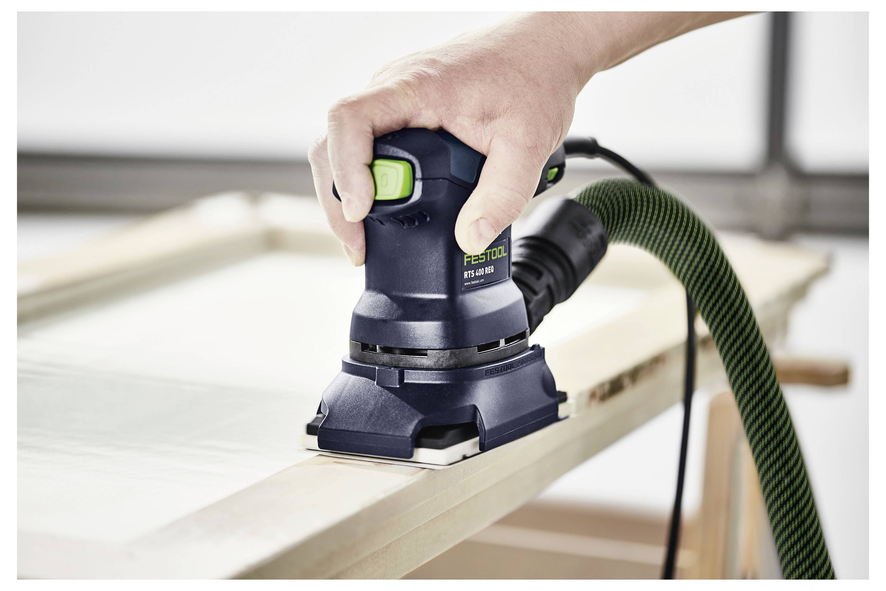 Una mano aziona una levigatrice elettrica su una superficie di legno, lisciando i bordi. La levigatrice è collegata a un tubo verde per l'estrazione della polvere.