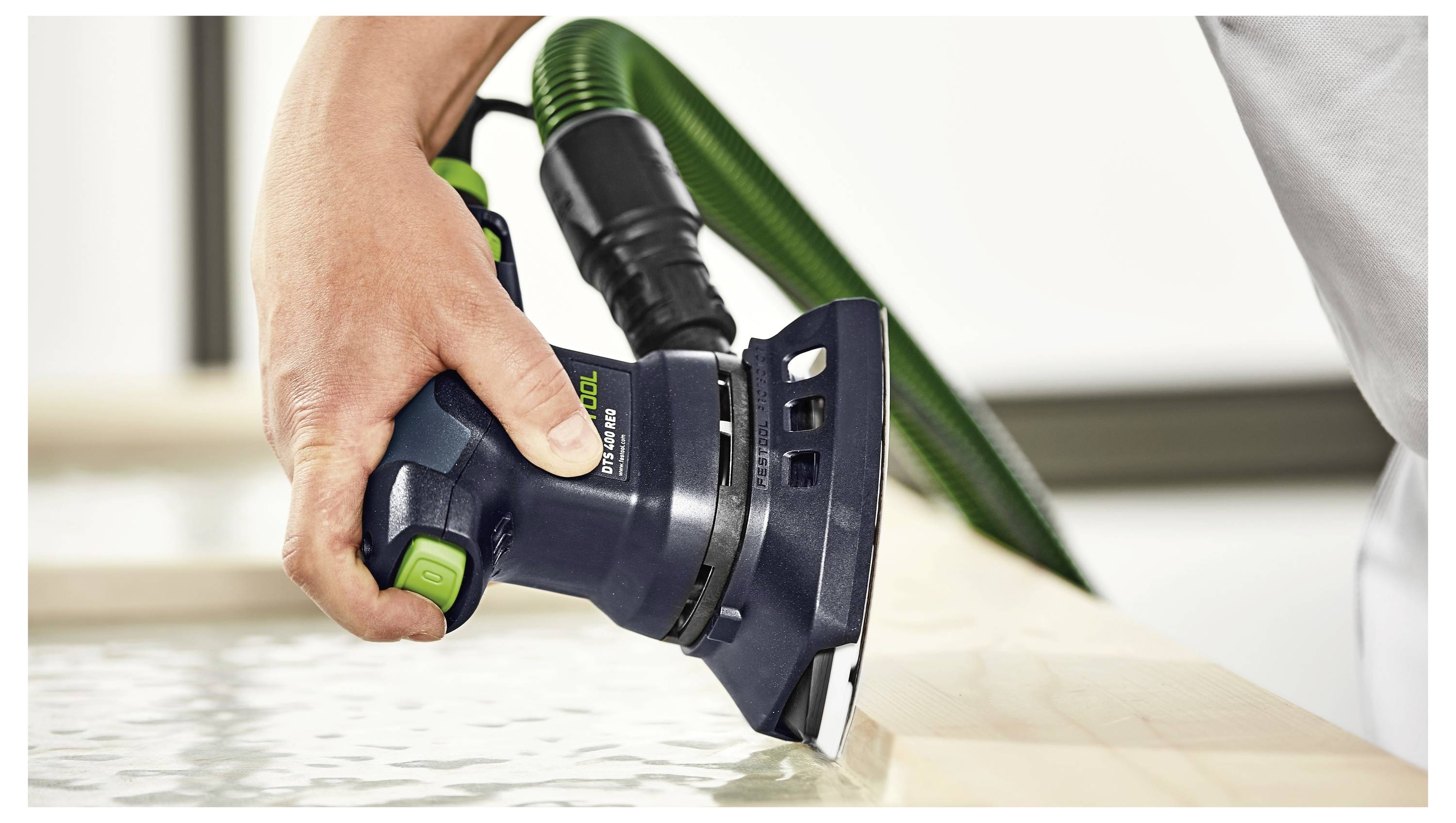 Una persona utilizza una levigatrice elettrica con un tubo di aspirazione verde per lisciare il bordo di una superficie in legno, concentrandosi sulla precisione e sulla finitura.