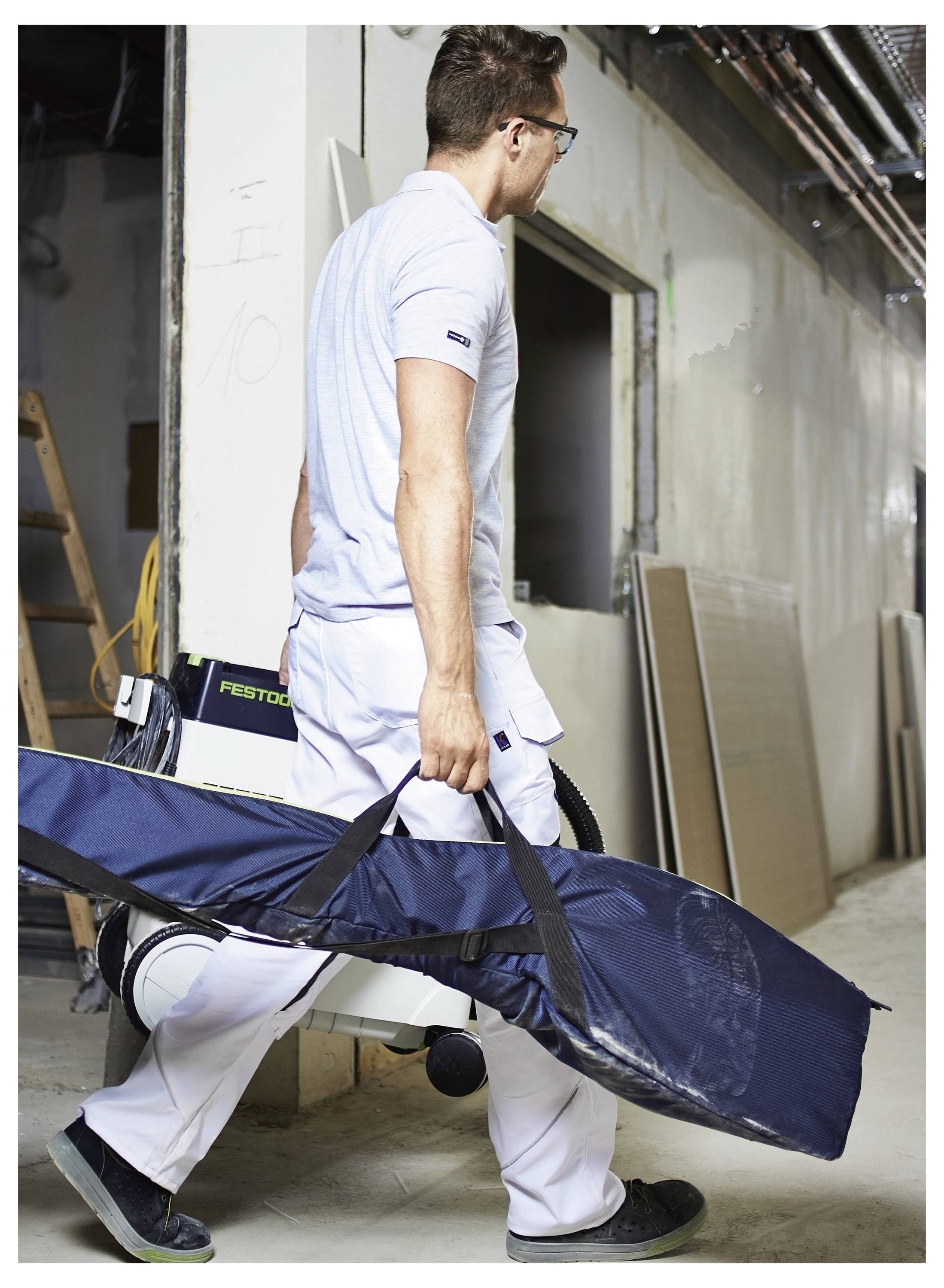 Una persona in un'ambientazione di laboratorio trasporta una custodia di trasporto blu e allungata. Indossa abbigliamento da lavoro bianco e occhiali, camminando accanto agli attrezzi.