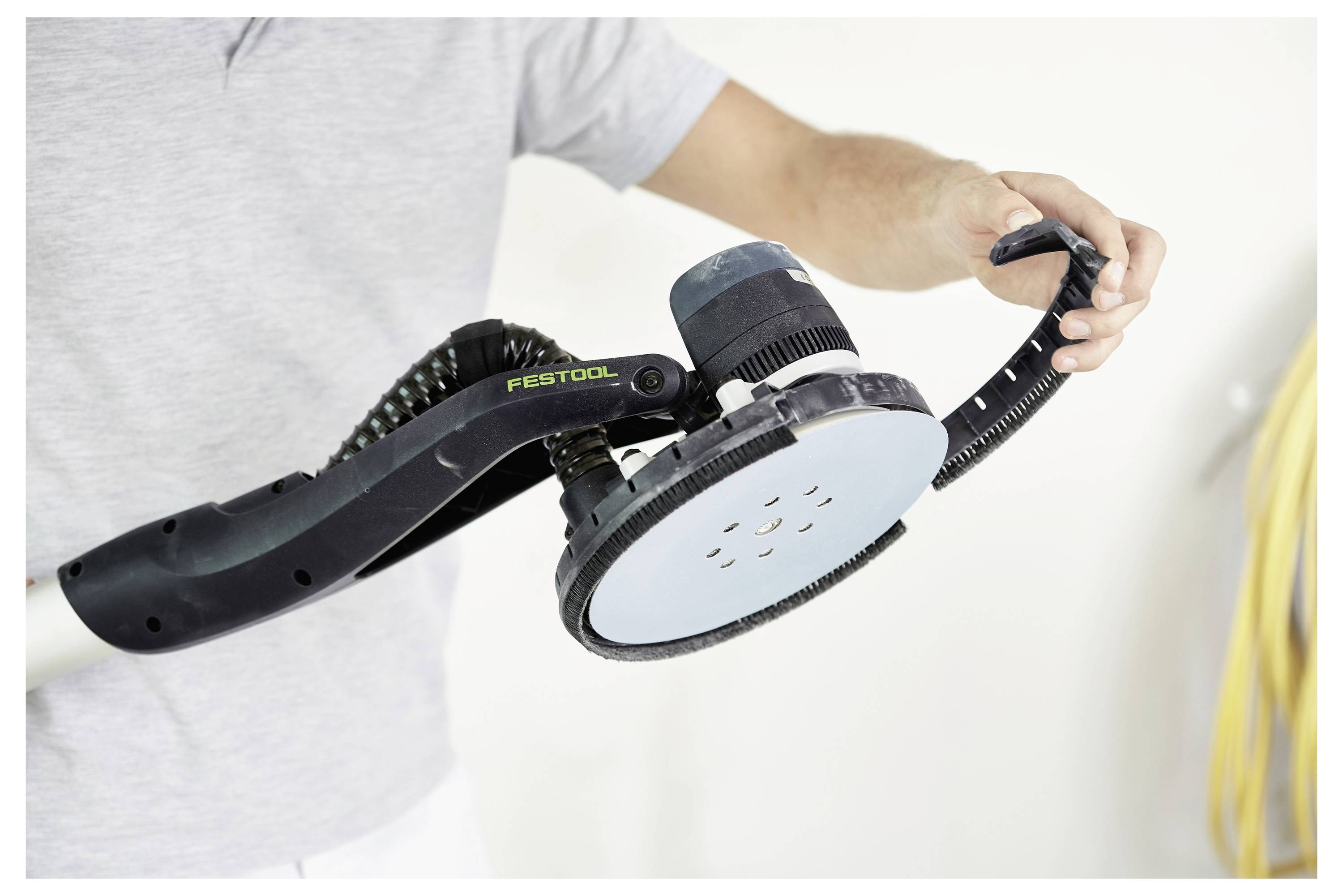Una persona tiene una levigatrice elettrica Festool nella mano destra, posizionata per l'uso. Lo sfondo è sfocato, con l'attenzione focalizzata sullo strumento.