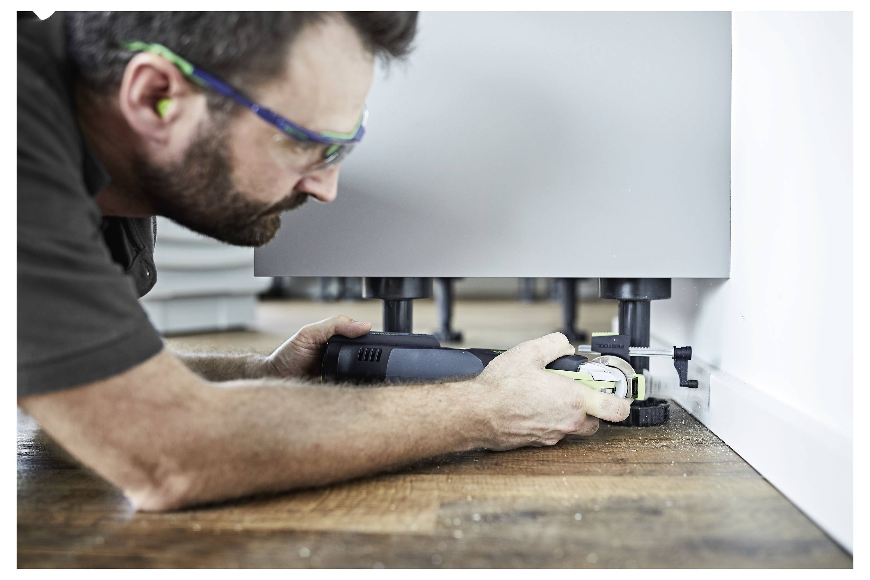 Uomo con occhiali protettivi che usa un attrezzo elettrico per regolare i piedi di un mobile su un pavimento in legno in una stanza.