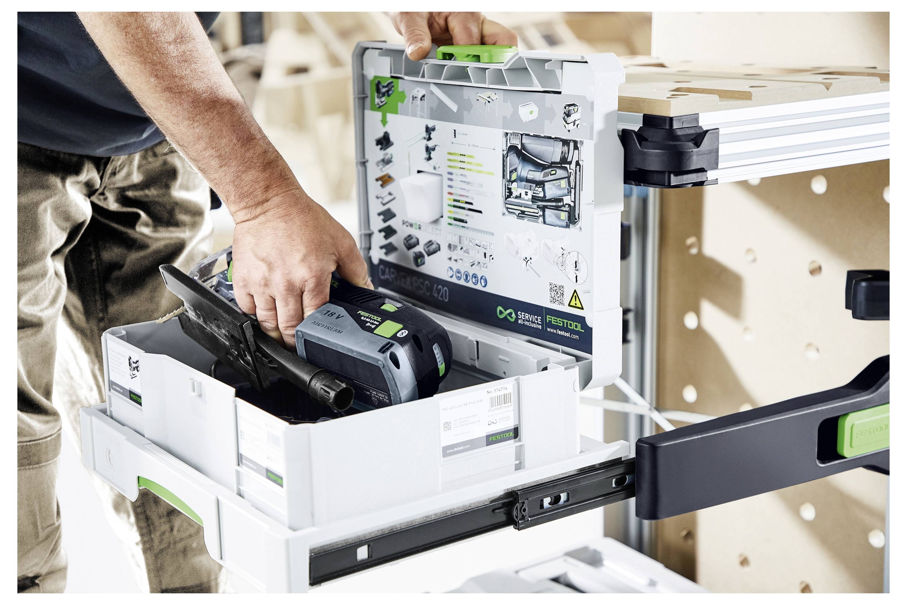 Una persona organizza un utensile elettrico all'interno di una scatola di stoccaggio etichettata in un laboratorio. La scatola si trova su uno scaffale estraibile collegato a un banco da lavoro.