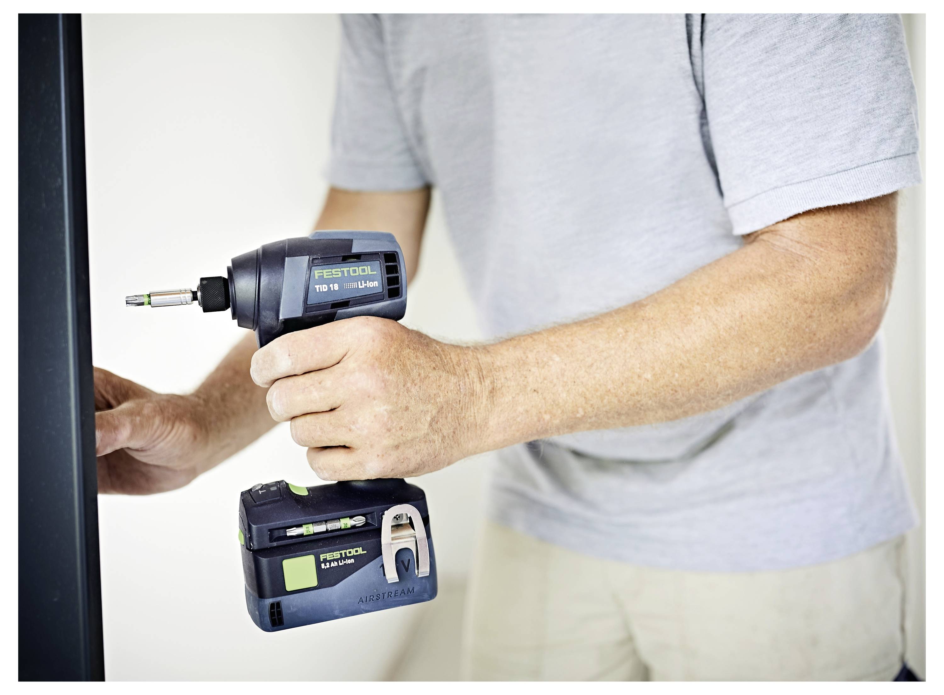 Una persona che indossa una maglietta grigia utilizza un trapano a percussione Festool su una superficie. L'attenzione è concentrata sul trapano in uso, mostrando il suo marchio e il design.