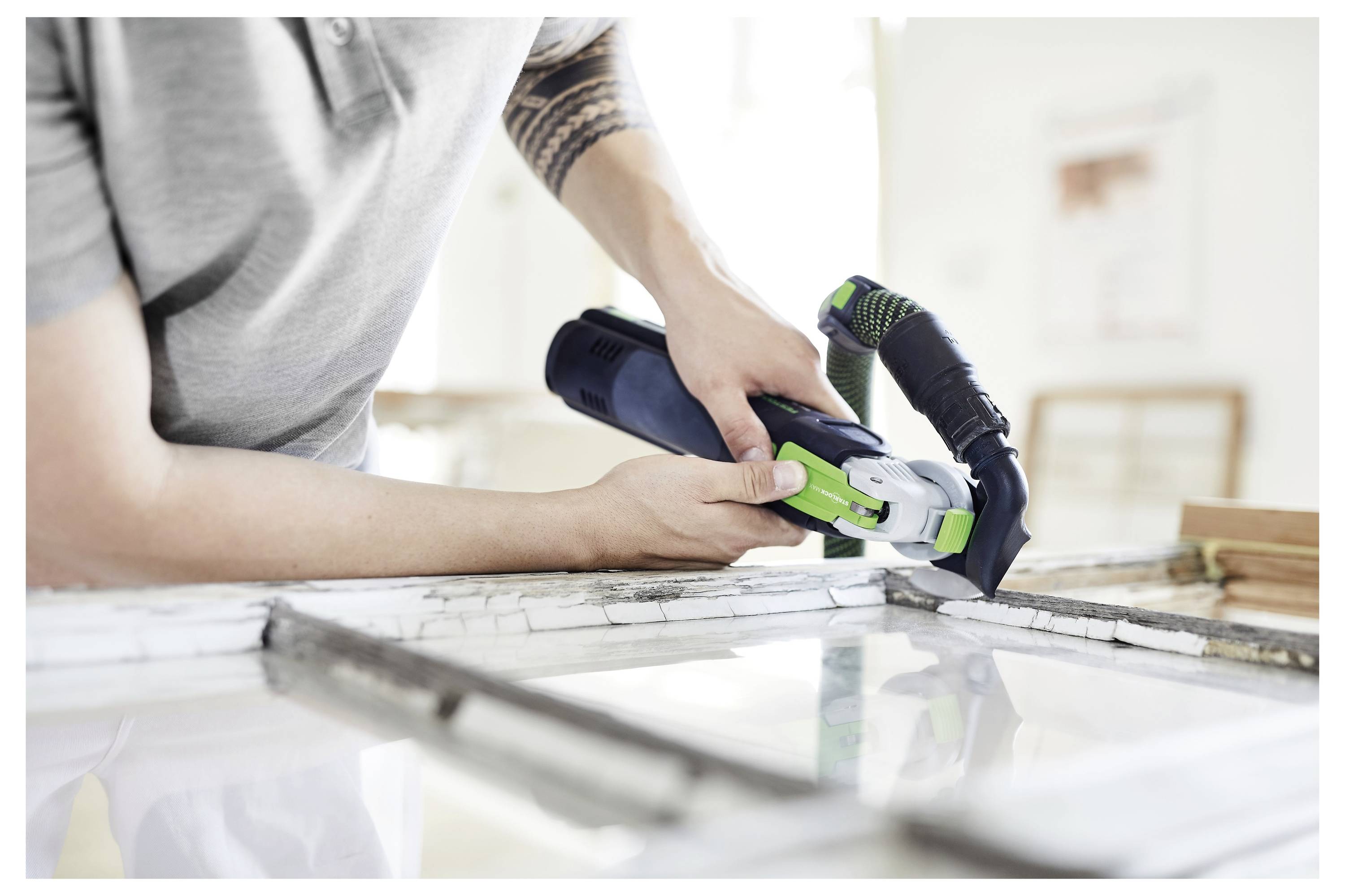 Una persona indossa una maglietta grigia e utilizza un attrezzo elettrico nero e verde per tagliare attraverso una cornice di finestra, mentre lavora a un progetto di ristrutturazione domestica.