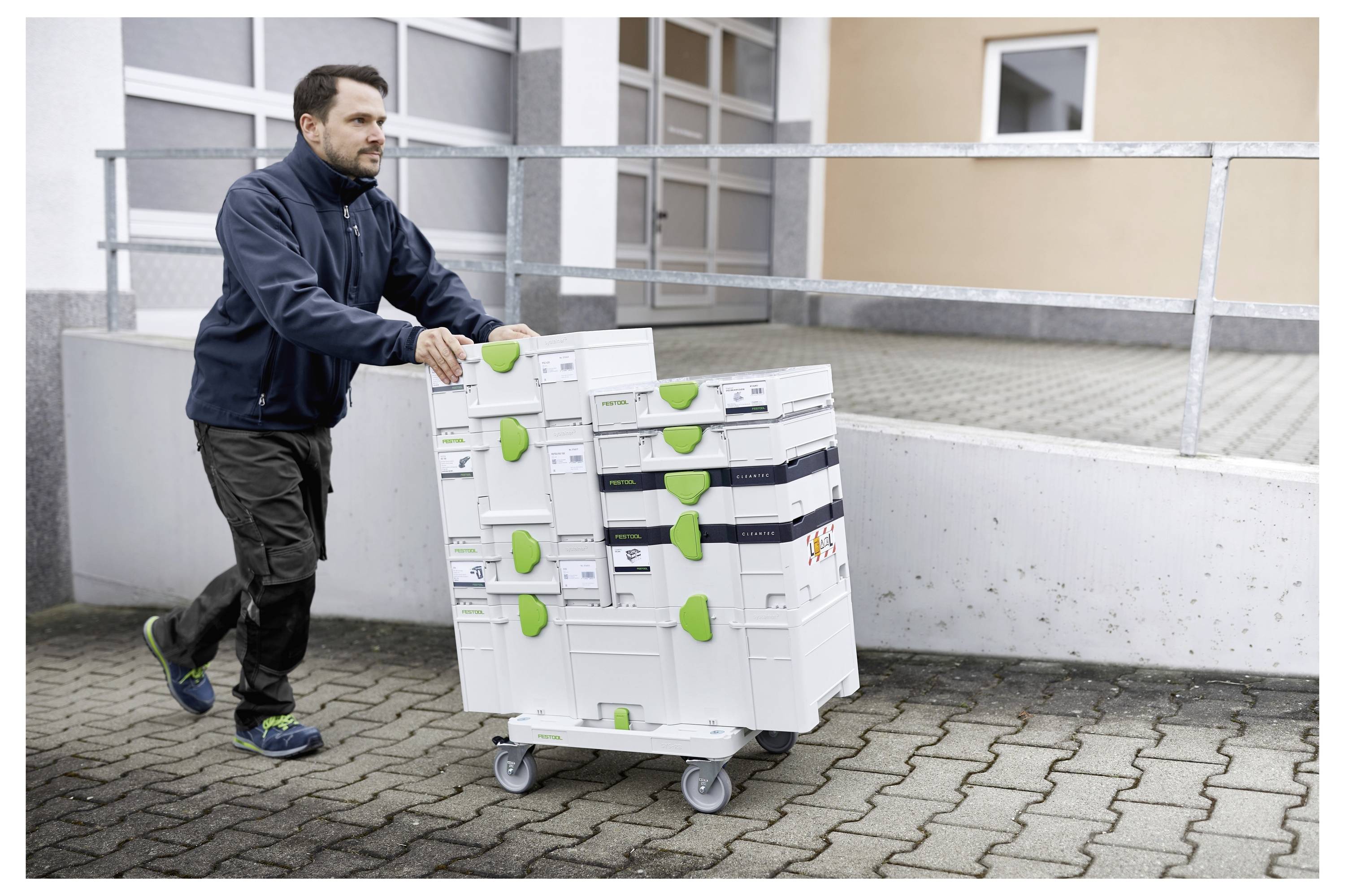 Un uomo che spinge un carrello con ruote carico di scatole di attrezzature su un'area pavimentata vicino a un edificio, suggerendo un trasporto o una consegna.