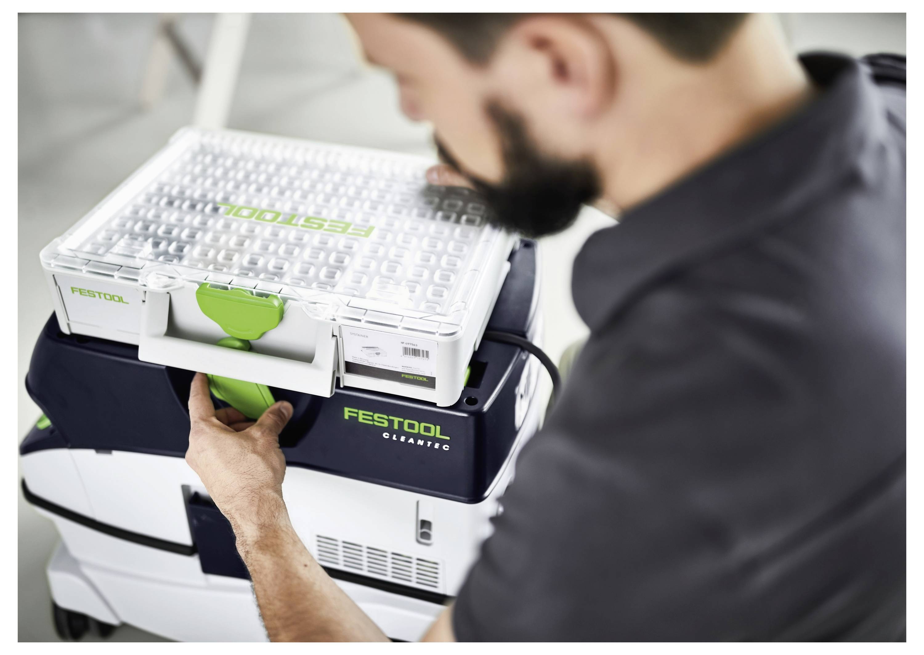 Una persona che posiziona una scatola di stoccaggio trasparente etichettata 'Festool' sopra un aspirapolvere Festool, indicando un'area di lavoro organizzata.