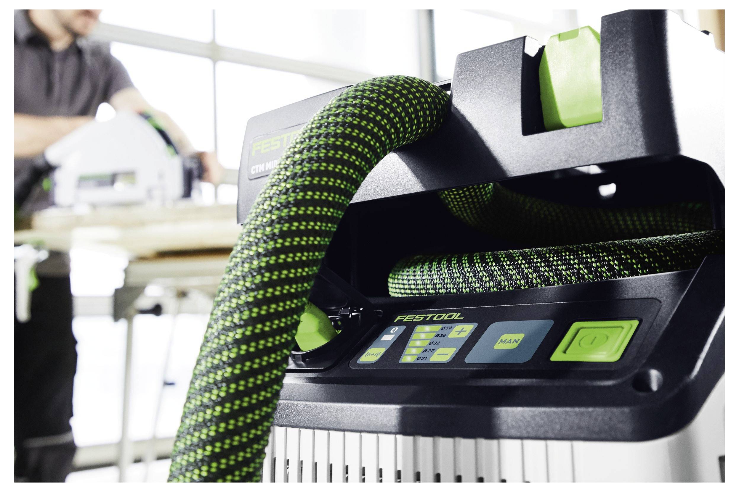 Un aspirapolvere con un tubo verde e nero è a fuoco, con una persona che lavora su una macchina in un'officina sullo sfondo.